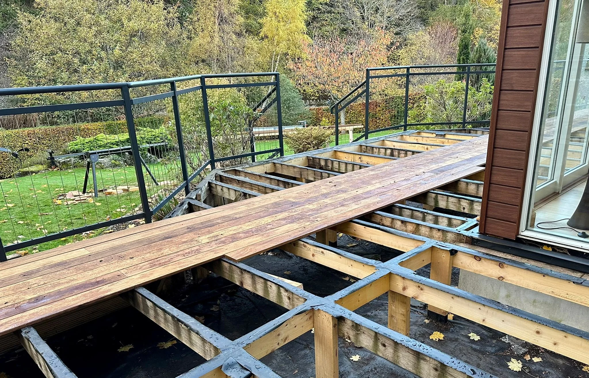 A wooden deck under construction on a house's backyard porch, with existing black metal safety railings and a view of a garden with trees and grass beyond.