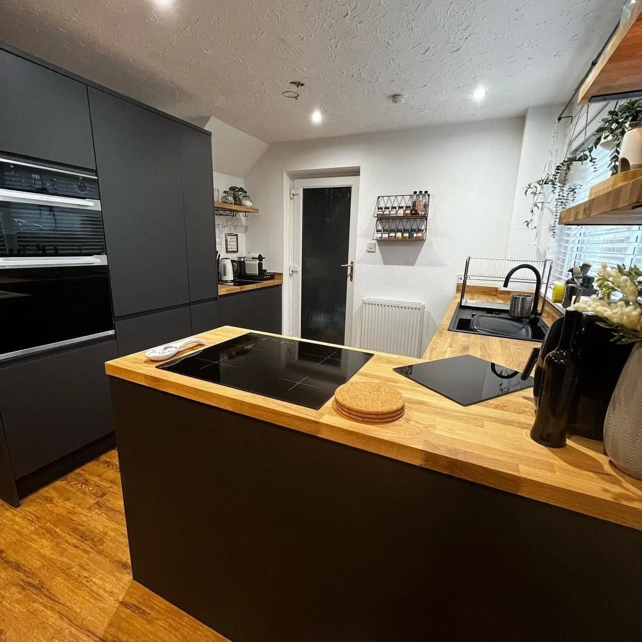 Modern kitchen with black cabinets, wooden countertops, and an electric stovetop on a central island. There is a black oven and microwave built into the cabinetry, a sink near a window, and various kitchen appliances and decor. The floor is wooden, and there's a door at the back with a chalkboard next to it.