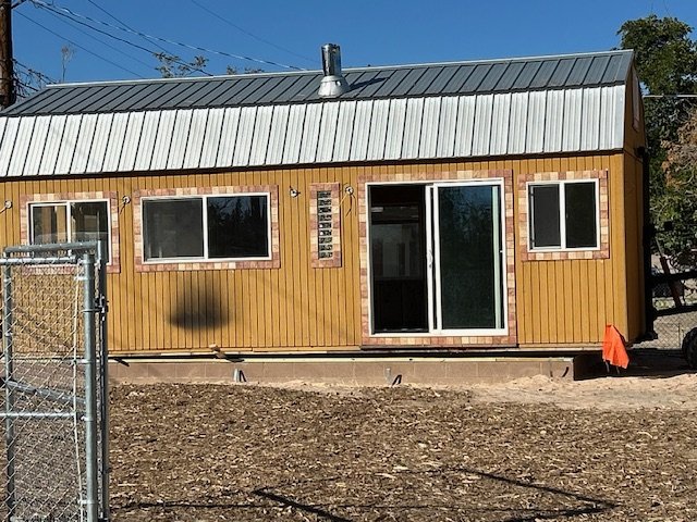Front view of a small wooden house with a metal roof, three windows, a sliding glass door, and a chain-link fence on the left side.