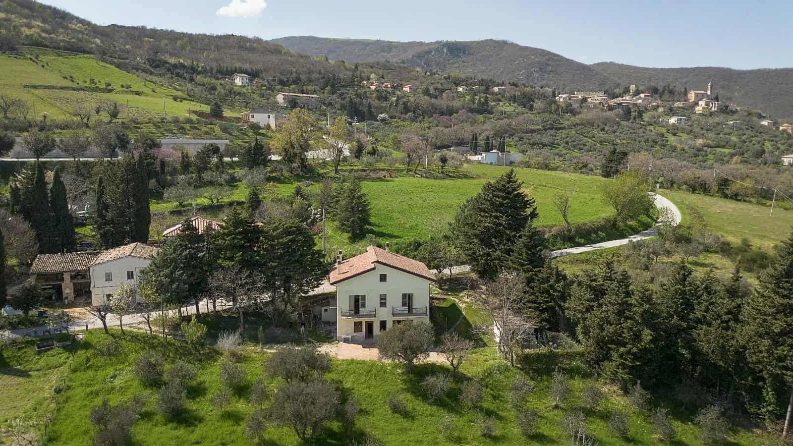 A countryside landscape featuring green rolling hills, scattered trees, and a few houses with tiled roofs, with mountains in the background.