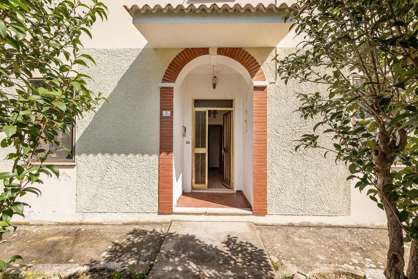 Front entrance of a house with a white textured exterior wall, brick archway, and a door with a screen door, surrounded by two trees.
