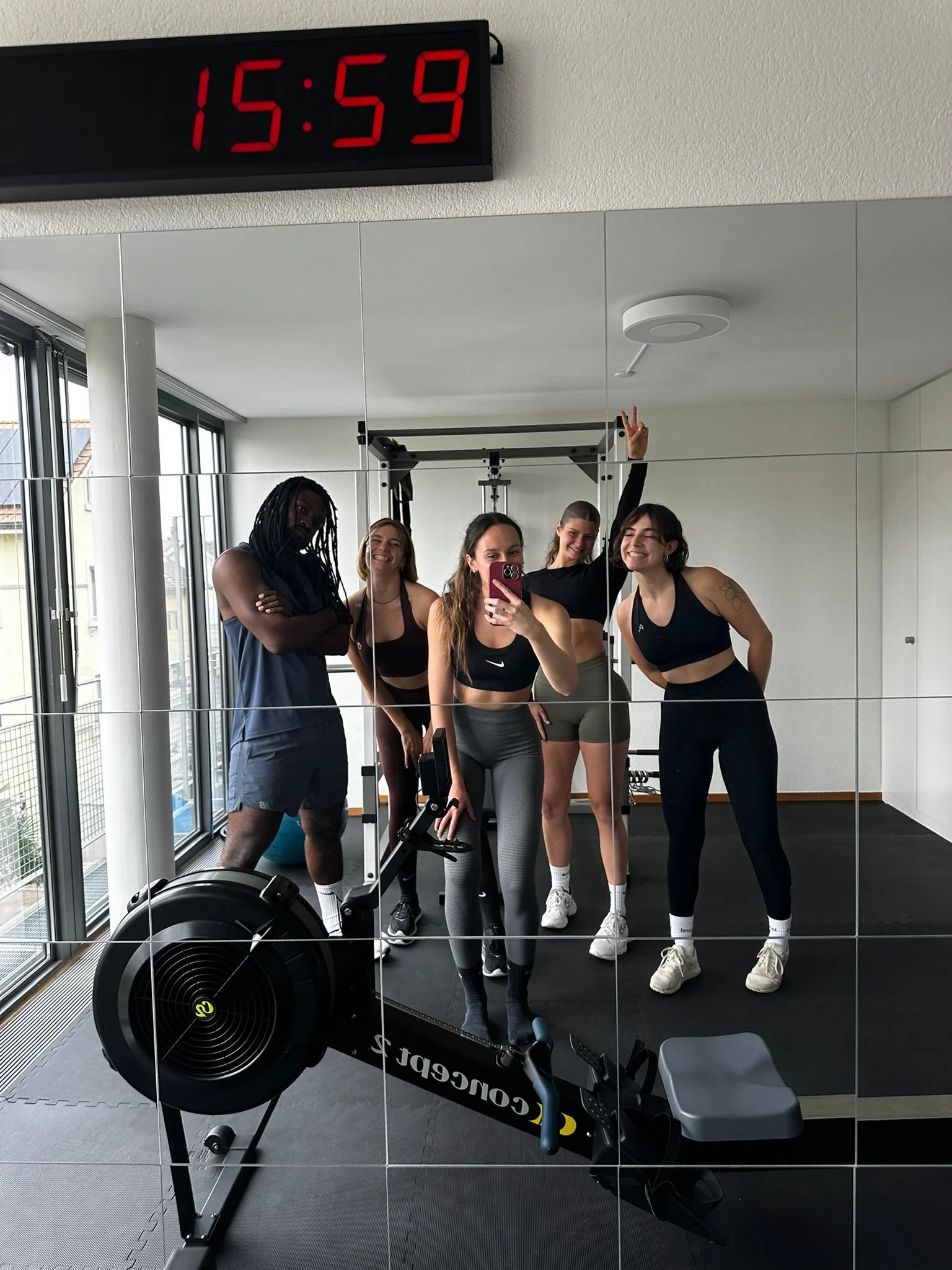 Un groupe de cinq femmes souriantes posent dans une salle de sport devant un miroir, avec un tapis de course à côté, portant des vêtements de sport.