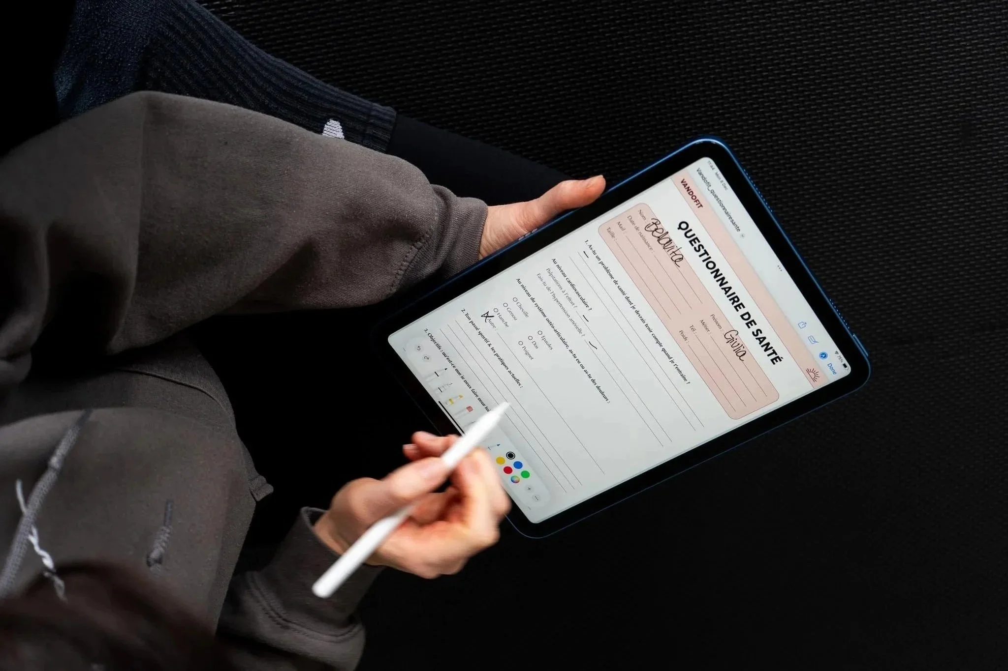 Une personne tient une tablette tactile avec un questionnaire de santé en français, intitulé 'QUESTIONNAIRE DE SANTÉ,' sur un fond noir. La personne utilise un stylet blanc pour répondre au questionnaire.