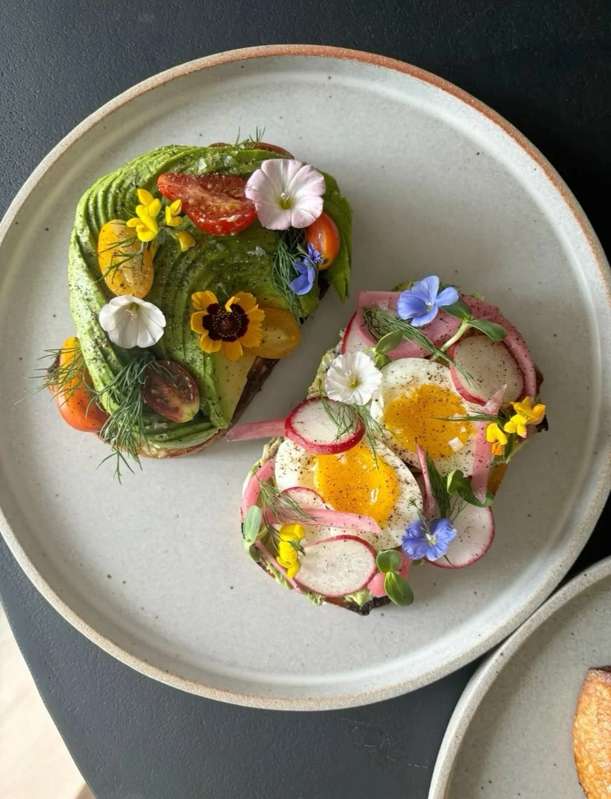 Deux toasts garnis d'œufs mollets, radis, bacon, herbes, micro-pousses et fleurs comestibles, présentés sur une assiette en céramique