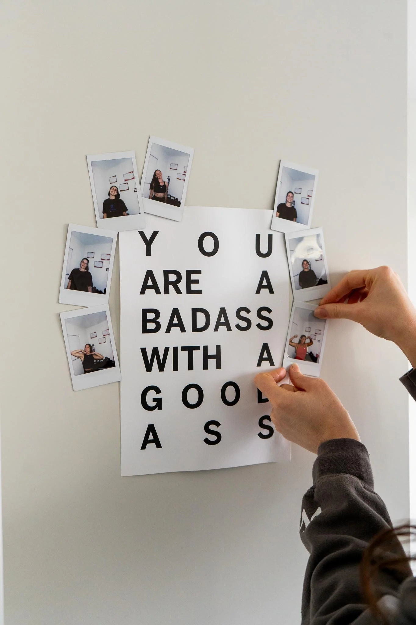 Une personne accroche des photos Polaroid autour d'une affiche avec le message 'YOU ARE A BADASS WITH A GOOD A$$' sur un mur blanc.