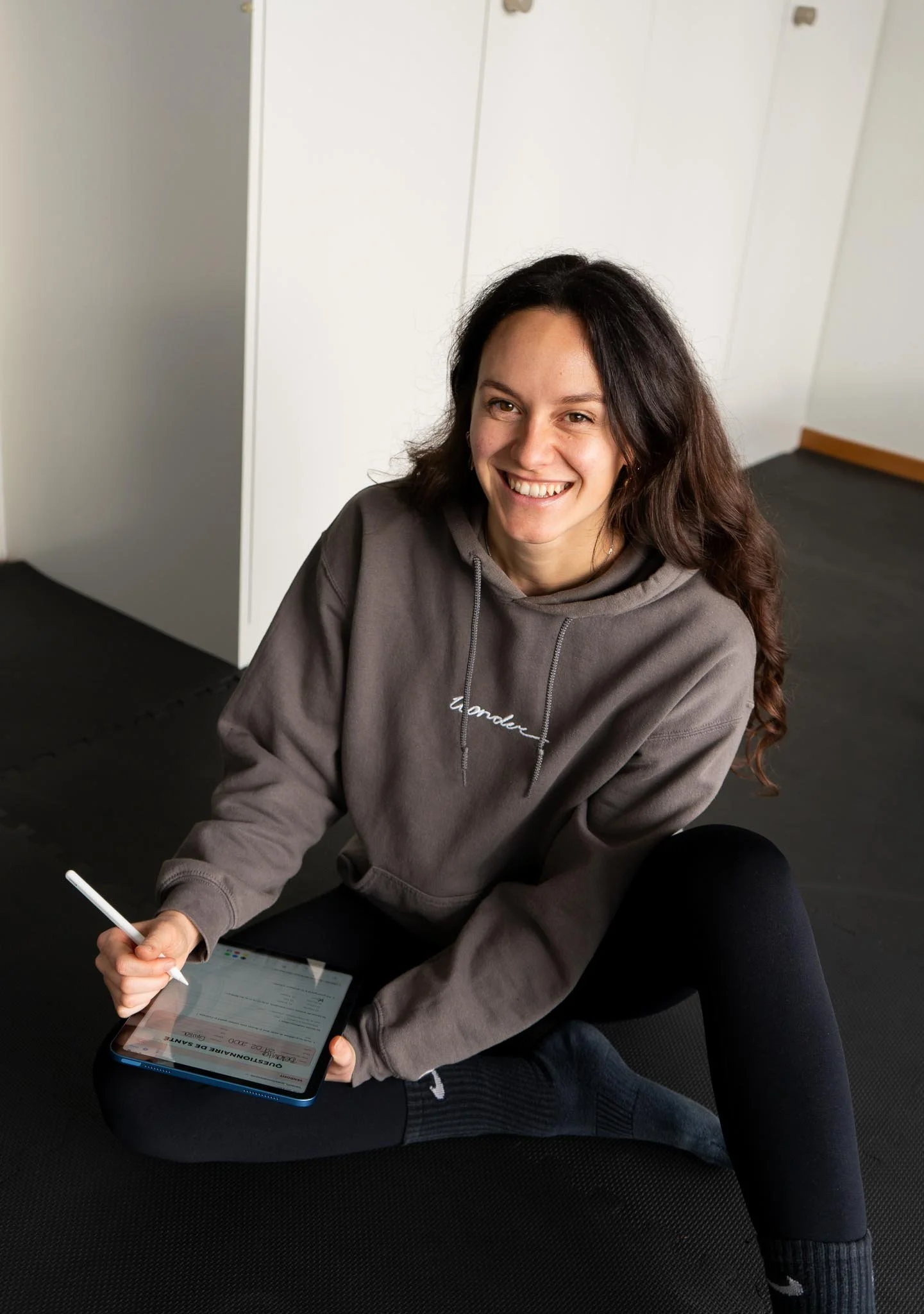 Une jeune femme souriante, assise sur un sol en caoutchouc, utilisant une tablette avec un stylet. Elle porte un sweat à capuche gris et des leggings noirs. L'environnement intérieur semble être une salle ou un studio de sport.