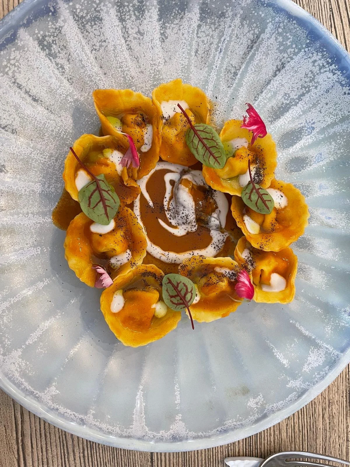 Assiette de raviolis de couleur orange garnis de feuilles vertes, de crème, de sauce brune, et de petites fleurs roses sur une assiette bleue avec fond en bois.