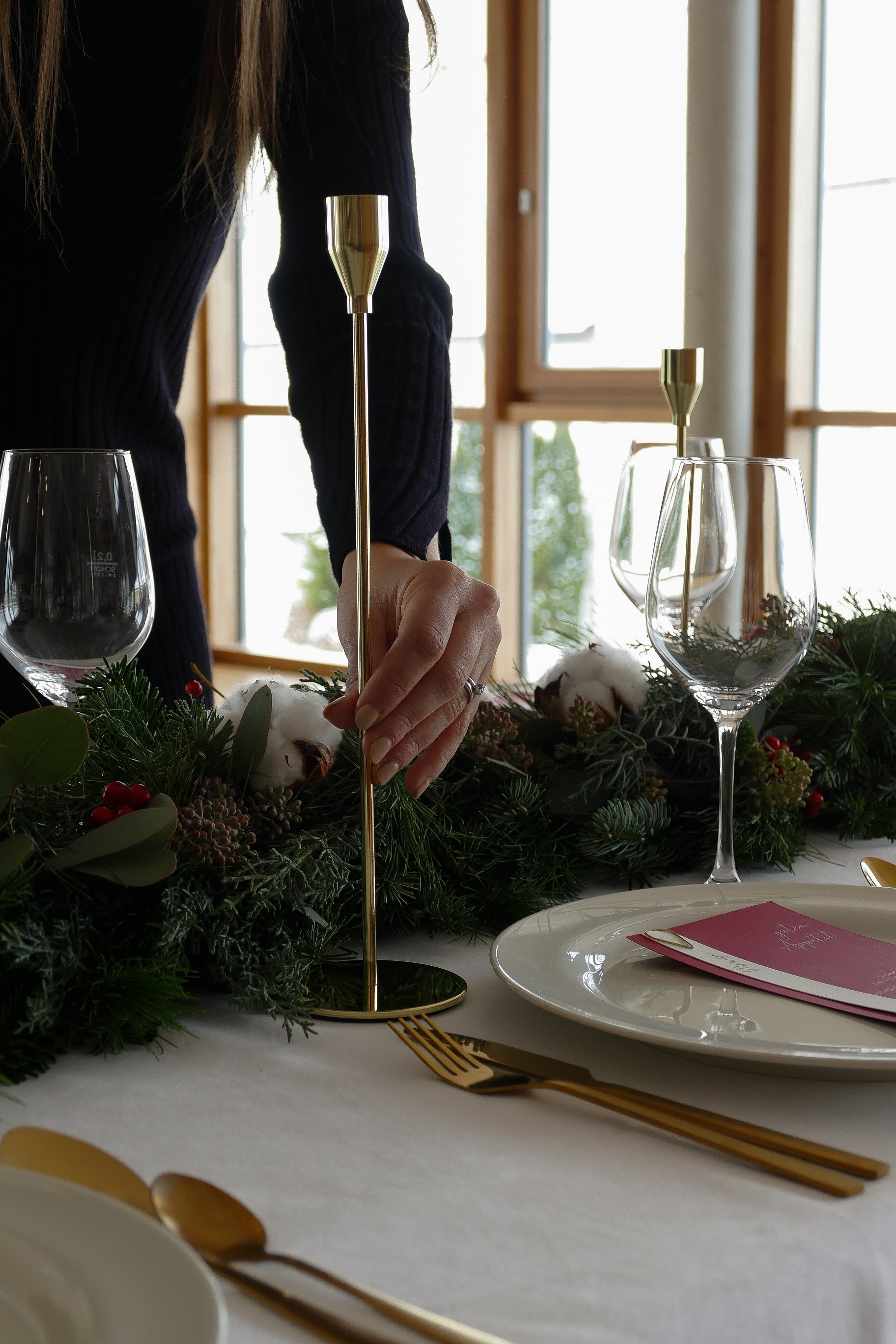 Eine Person dekoriert einen festlich gedeckten Esstisch mit einer goldenen Kerzenhalterung, Weihnachtsdeko mit Tannenzweigen, Baumwolle und Beeren, Gläsern und Geschirr.