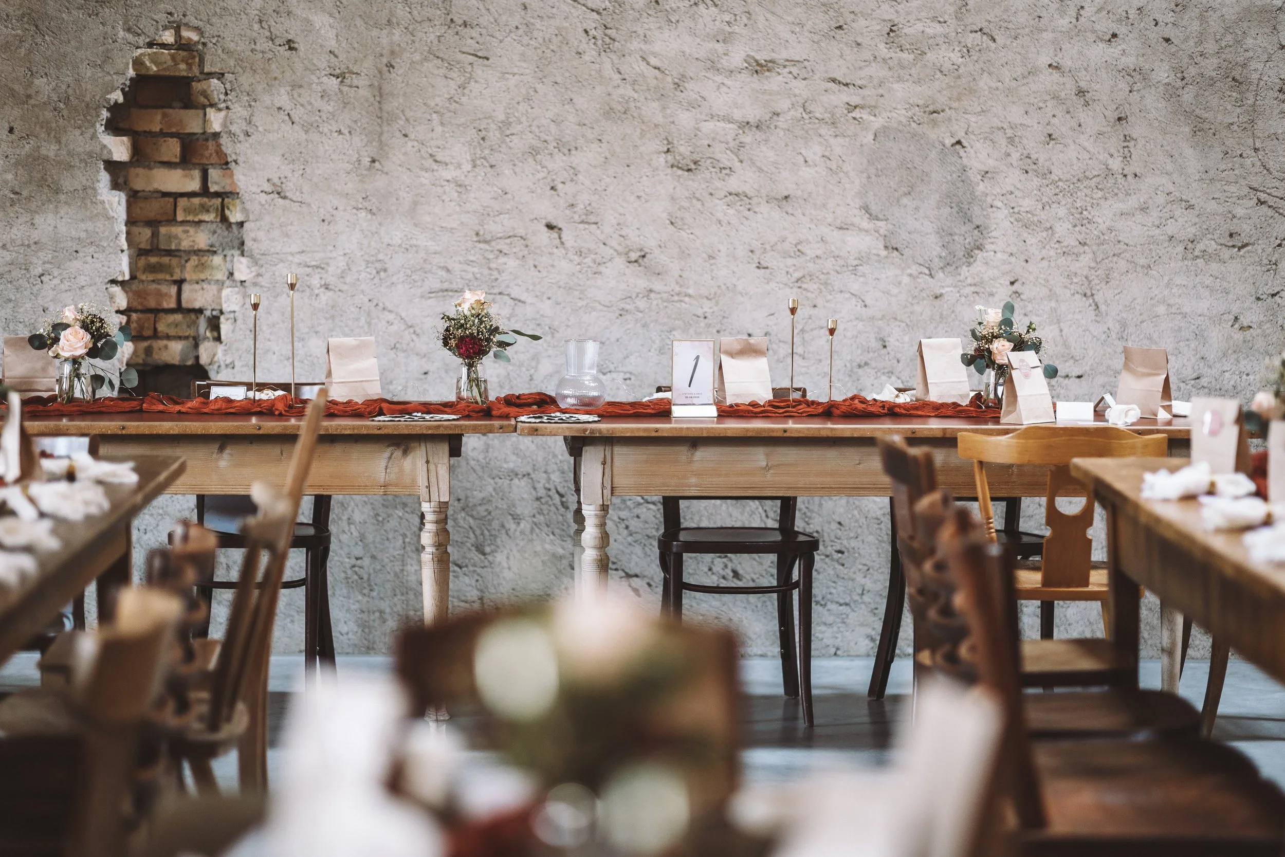 Eine dekorierte Festtafel in einer rustikalen, offenen Location mit einer verputzten Steinwand im Hintergrund, gedeckt mit Blumen, goldenen Kerzenhaltern, sowie rostfarbenen Tischläufern.