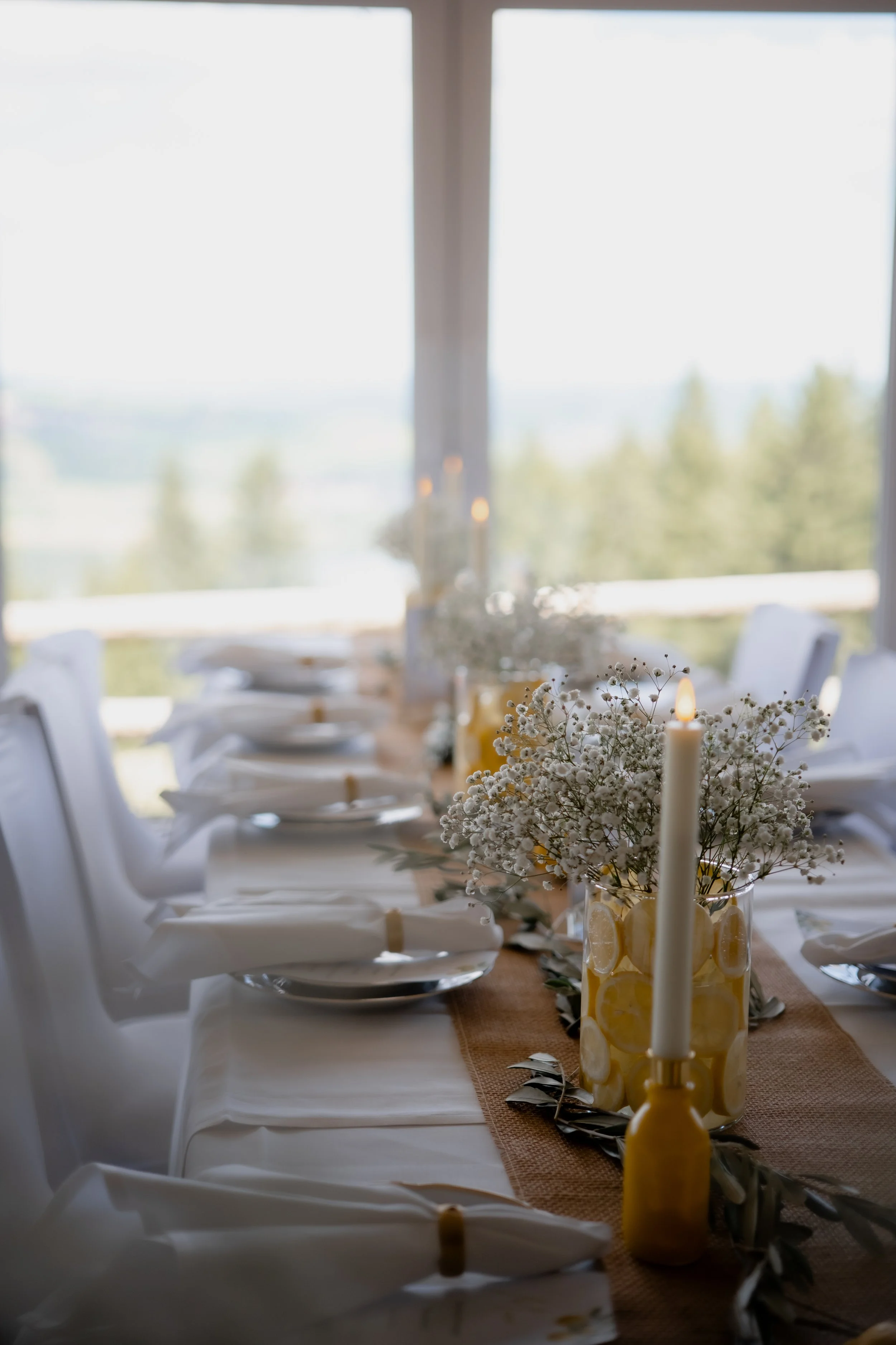 Eine schön gedeckte Festtafel mit weißen Tellern, Besteck, weißen Stoffservietten mit goldenen Serviettenringen und Kerzen in Glaszylindern, mit Blumendekoration aus weißen Blumen, vor einer großen Fensterfront mit Blick ins Grüne.