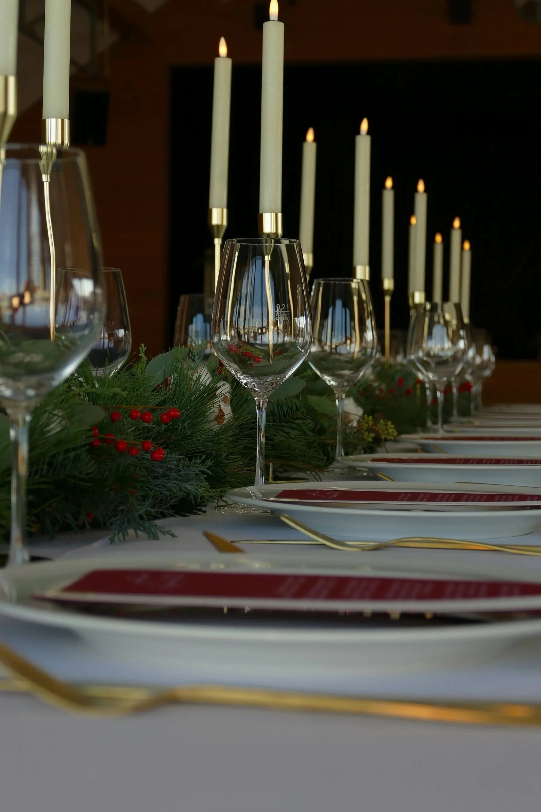 Festlich gedeckter Tisch mit weißen Tellern,Weingläsern, goldenen Bestecken und Kerzen auf einer Weihnachtsdekoration aus Tanne und roten Beeren.