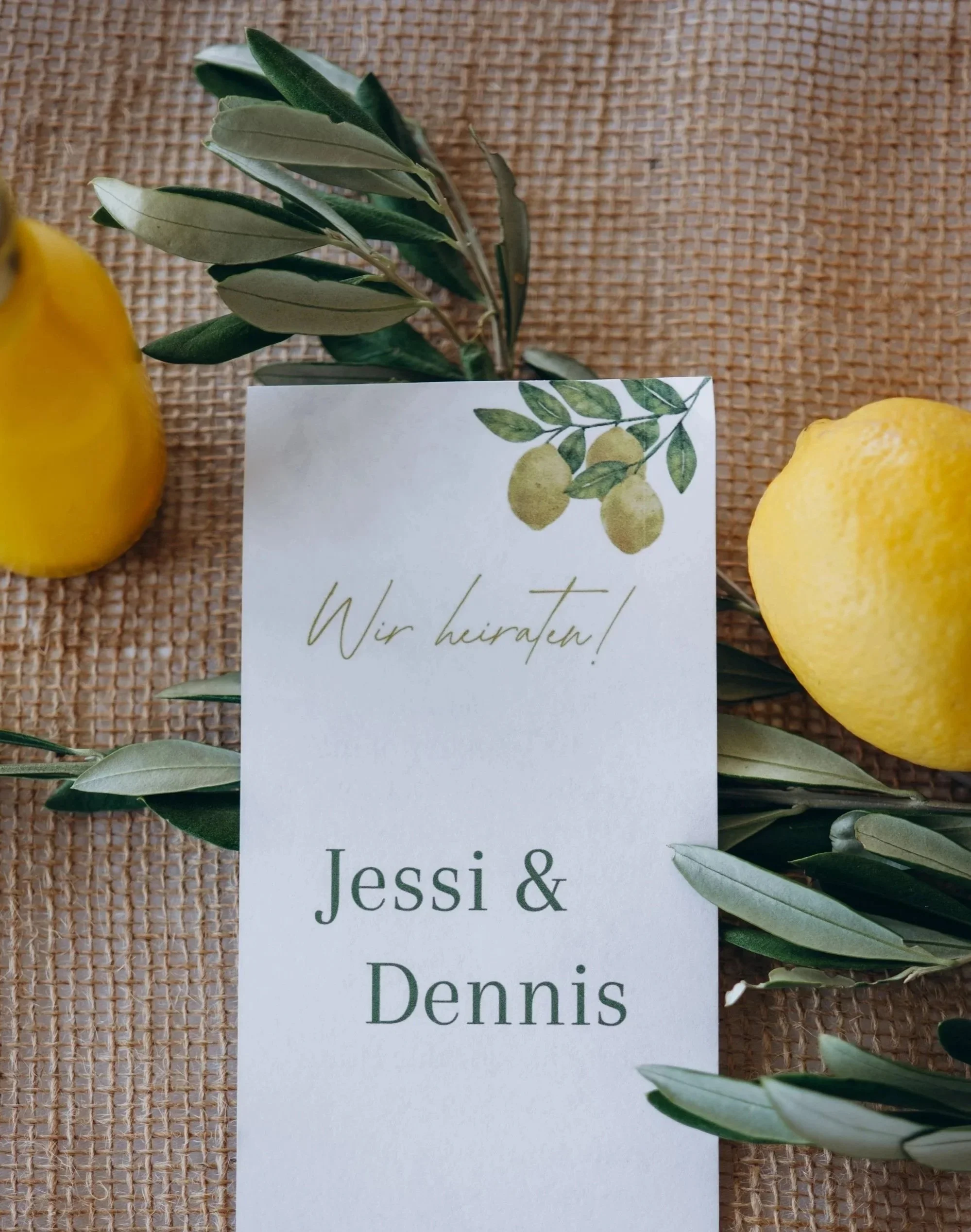 Eine Einladungskarte mit Hochzeitsgruß, umgeben von Zitronen und Olivenzweigen auf einem Jute-Tischläufer.