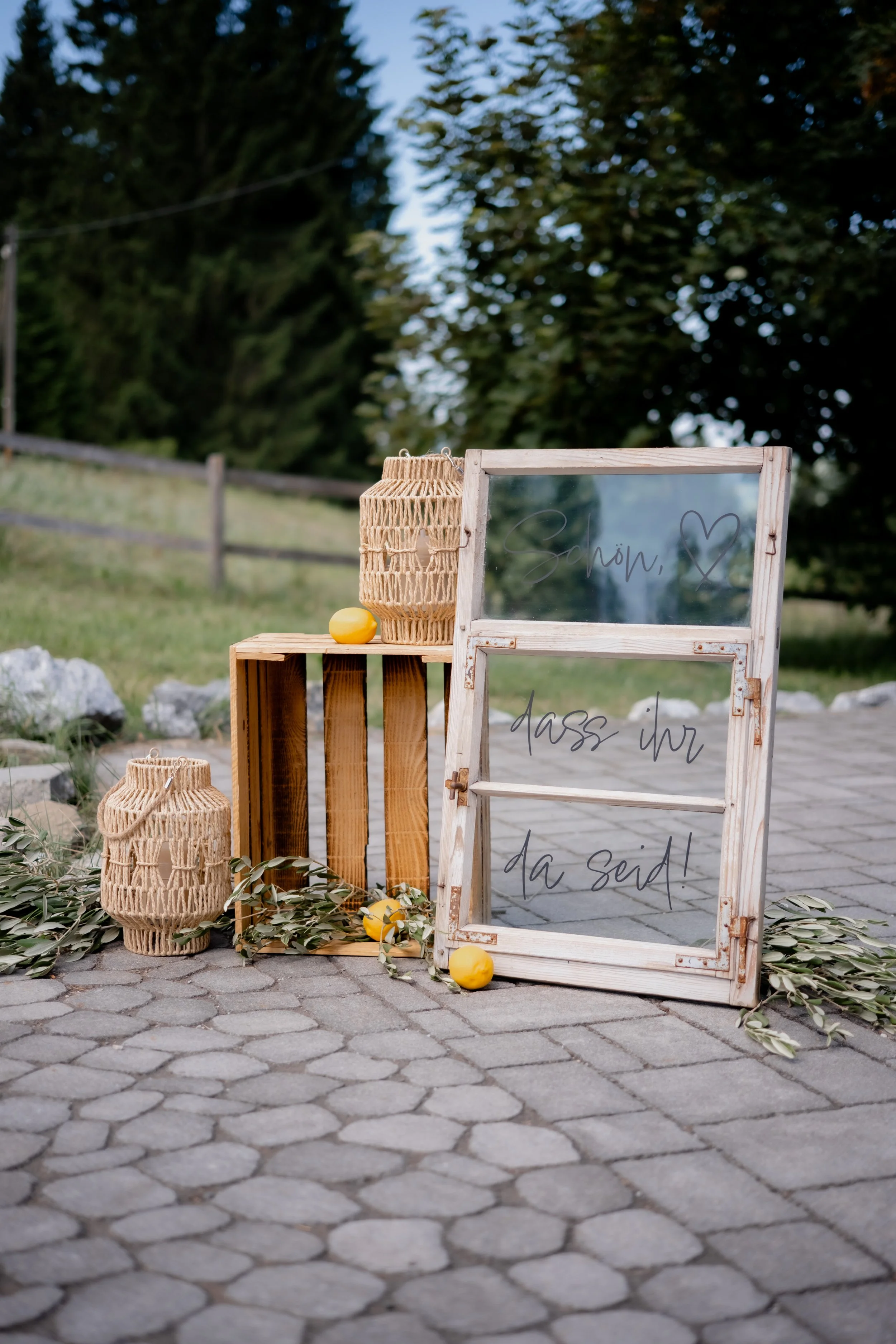 Dekoratives, rustikales Willkommensschild auf gepflastertem Boden, umgeben von Papierlaternen, Zitronen und grünen Olivenzweigen, mit Bäumen im Hintergrund.