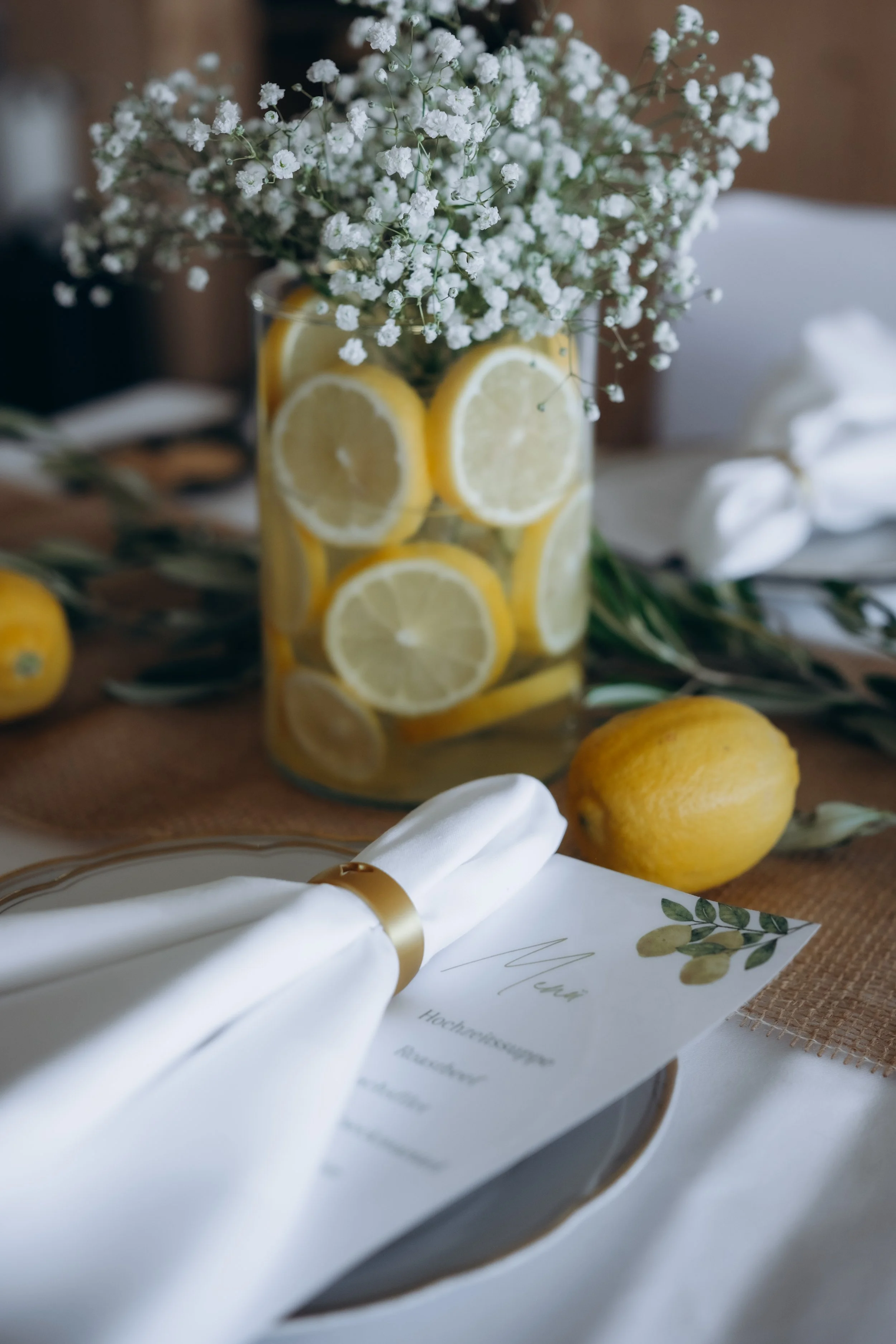 Mediterrane Tischdeko einer Hochzeit, Blumenstrauß mit weißen Blumen und Zitronen in Glaszylinder auf gedecktem Tisch mit weißer Tischdecke und Jute-Tischläufer, sowie weiße Serviette mit goldenem Serviettenring und einer Menükarte auf dem Teller.