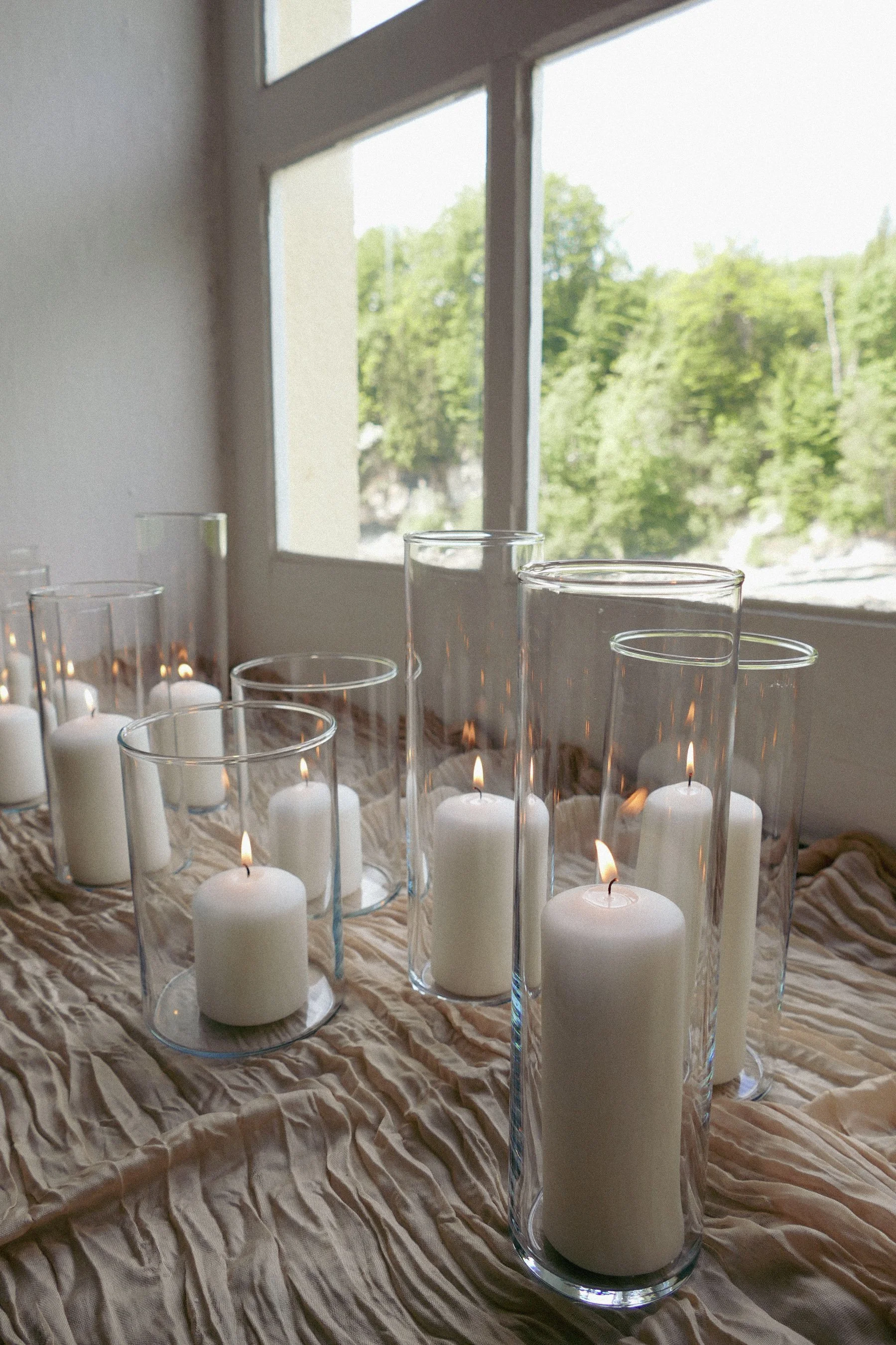 Weiße Kerzen in hohen transparenten Glaszylindern auf beigem Läufer vor einem großen Fenster mit Blick auf grüne Bäume.