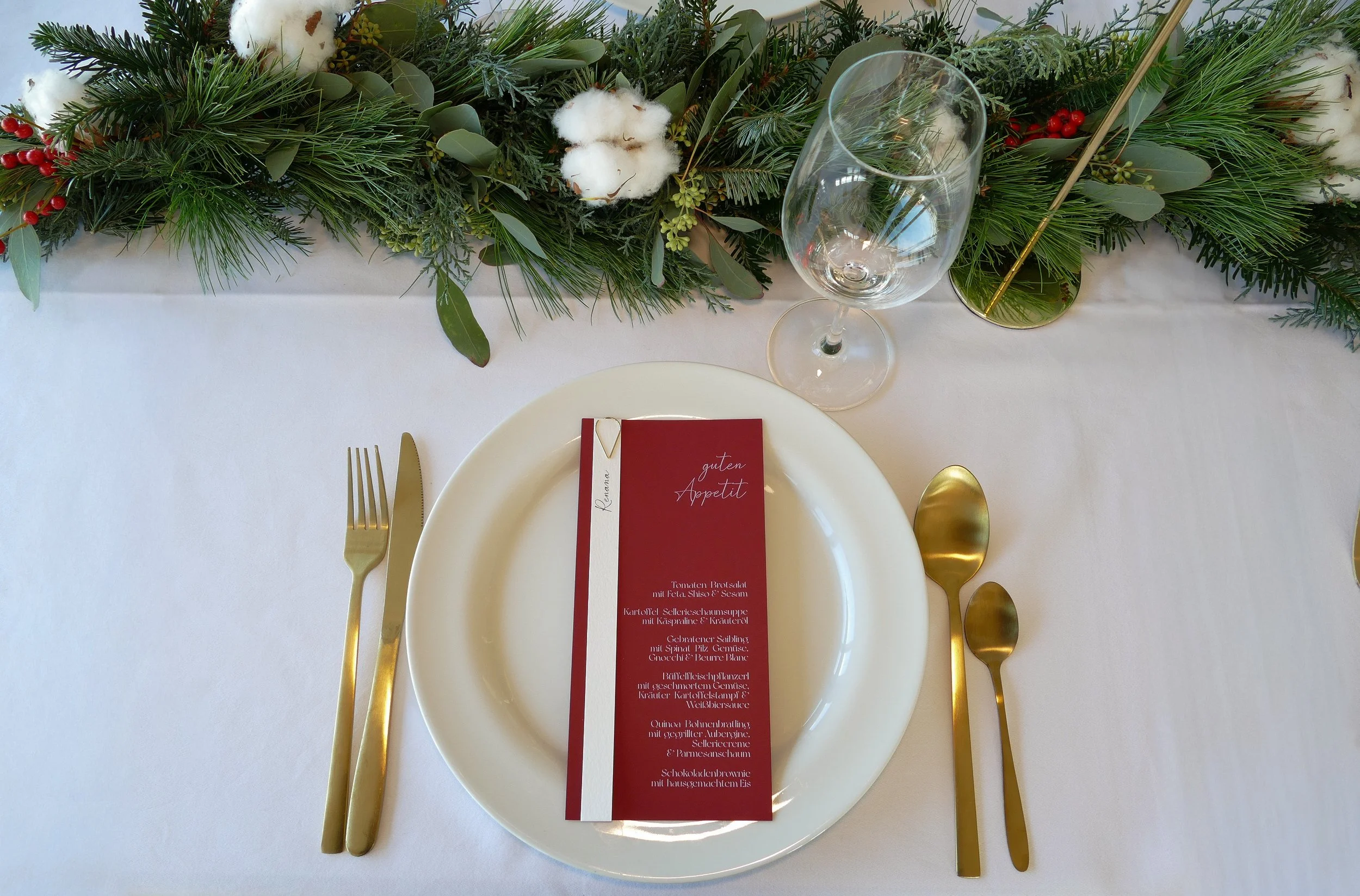 Weihnachtliche Tischdekoration mit einem weißen Teller, goldenem Besteck, einem leeren Weinglas und einer grünen Tafelkerze auf einem weißen Tischtuch, darüber eine dekorative Anordnung aus Tannenzweigen, Baumwollzweigen, roten Beeren und Eukalyptusblättern.