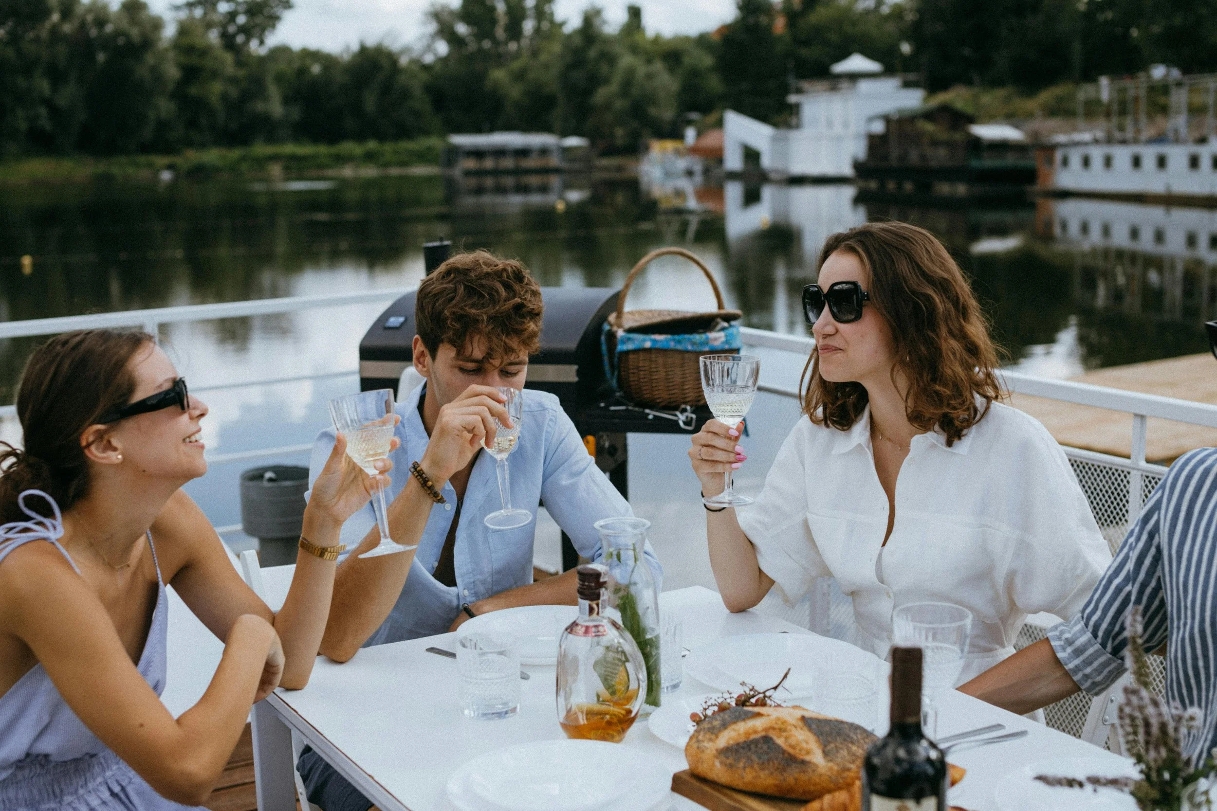 Group of four people enjoying drinks and food on a boat deck, with a scenic waterway and boats in the background.