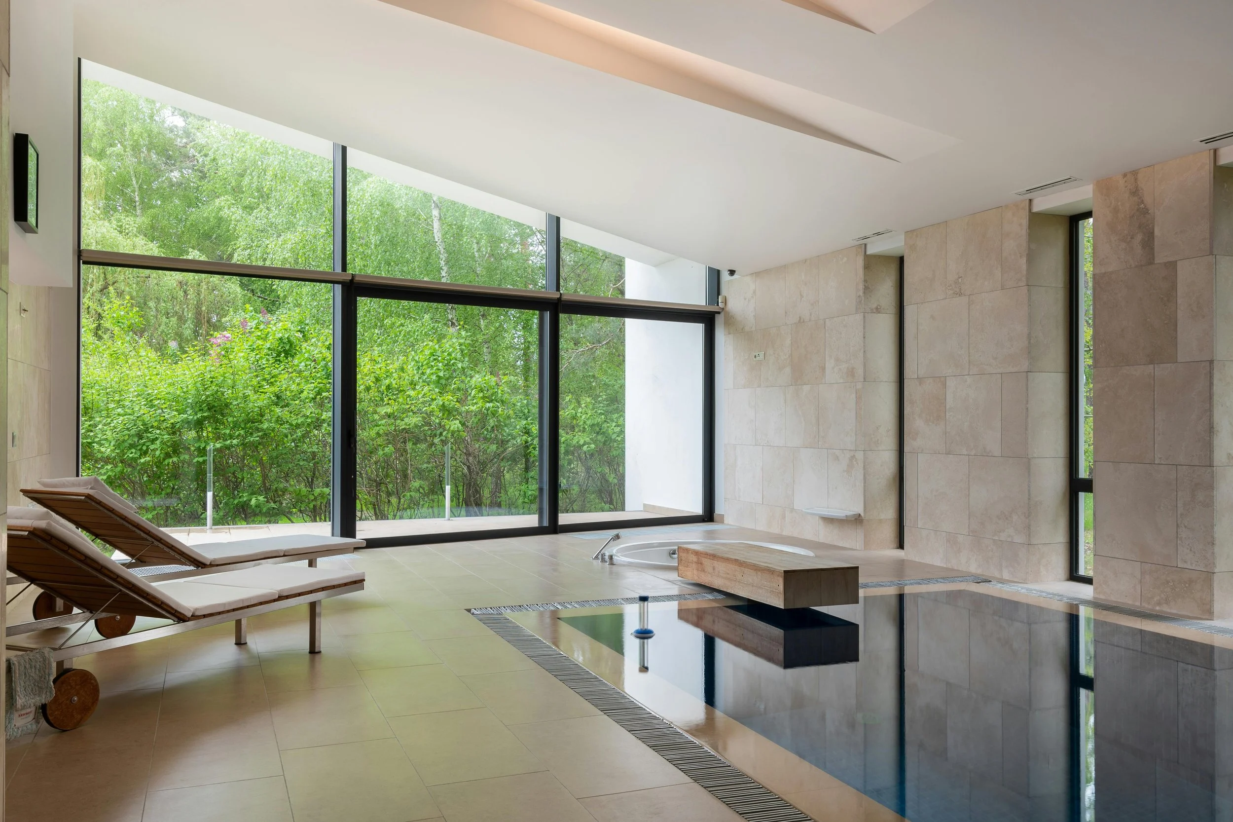 Indoor pool area with beige tiled floor and walls, large glass windows showing green trees outside, two lounge chairs, and a small hot tub.