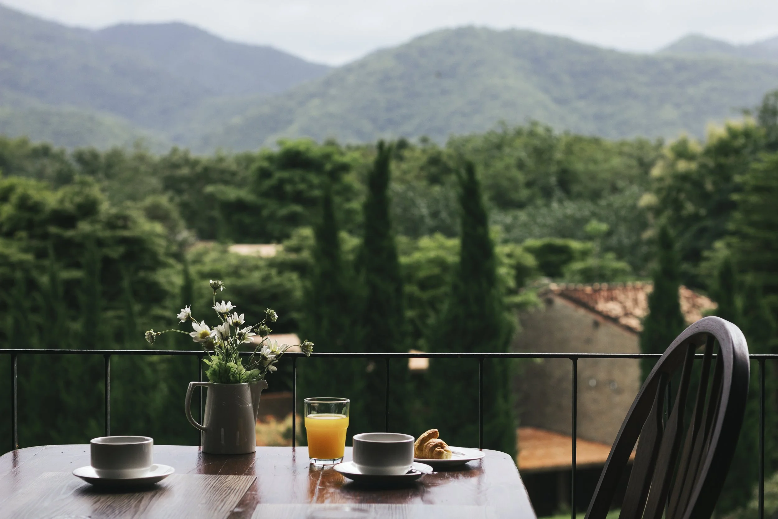 A breakfast table with a vase of white flowers, a glass of orange juice, two white cups, and a croissant, set on a balcony with a lush green landscape and mountains in the background.