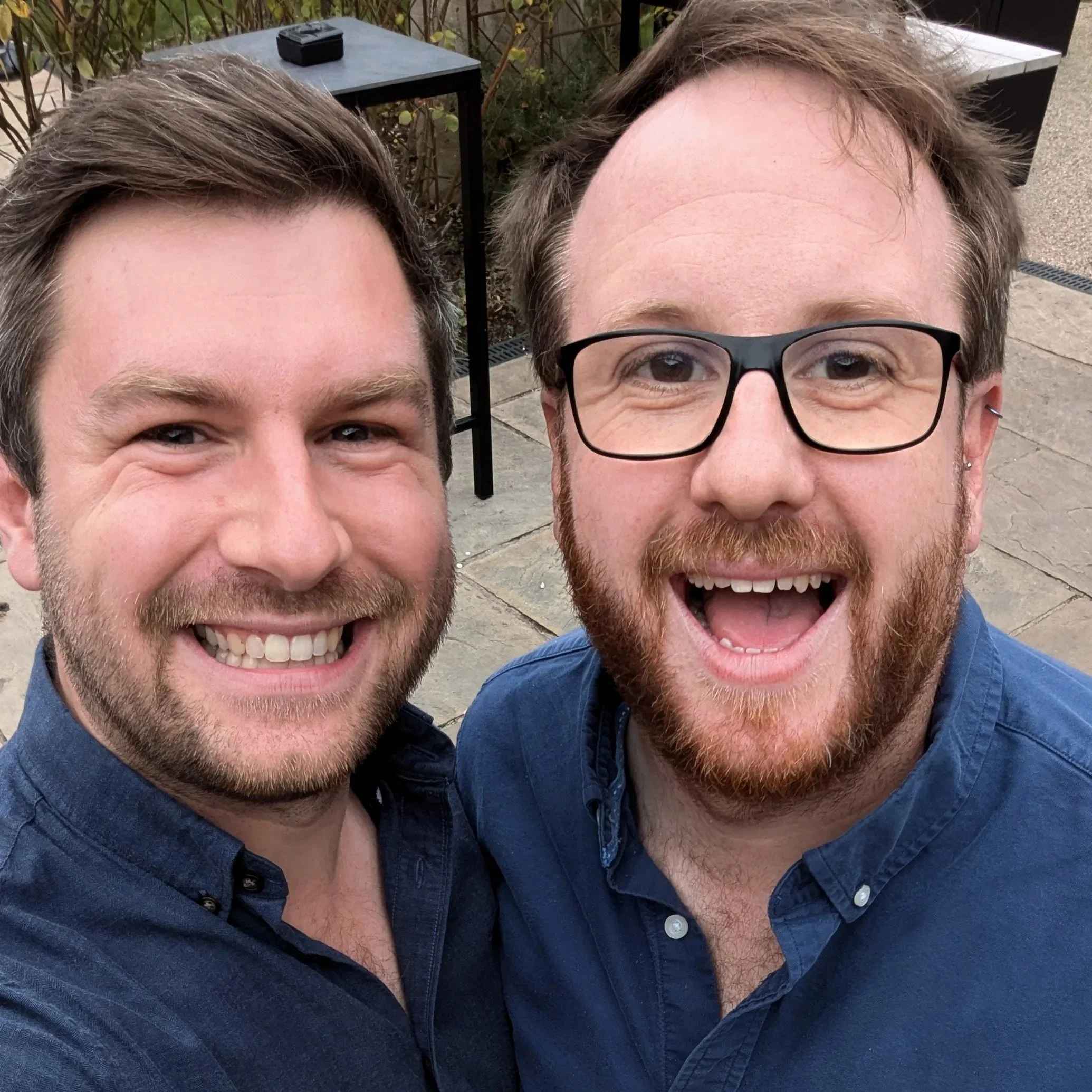 Two men smiling and taking a selfie outdoors in a casual setting, wearing blue shirts.