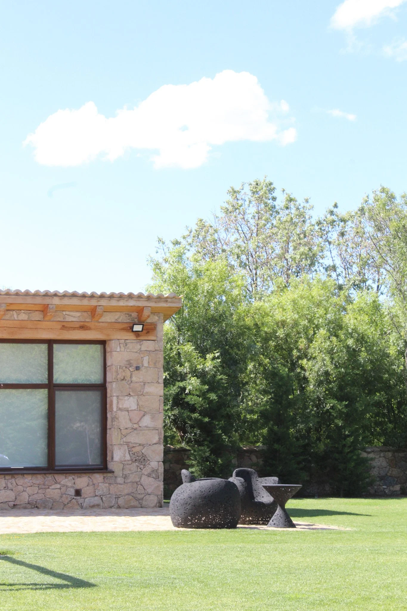 Exterior de una casa con muro de piedra, ventana grande con marco de madera y vista a un jardín con esculturas negras y árboles verdes bajo un cielo azul con nubes blancas.
