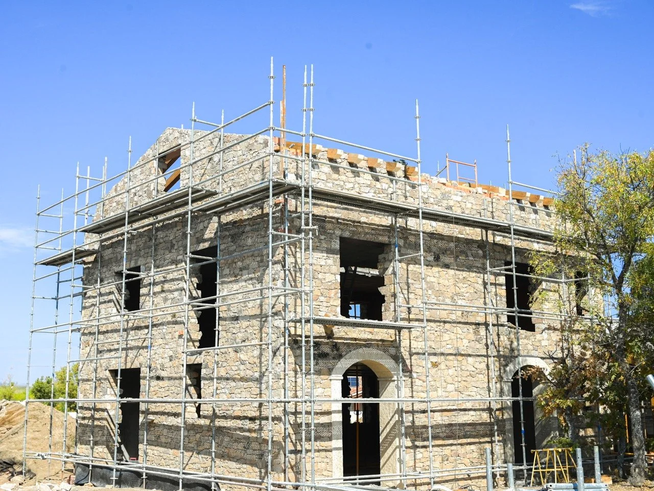 Edificio en construcción rodeado de andamios de metal, hecho de piedra, con ventanas y puertas enmarcadas en piedra, bajo un cielo azul, con árboles cercanos.