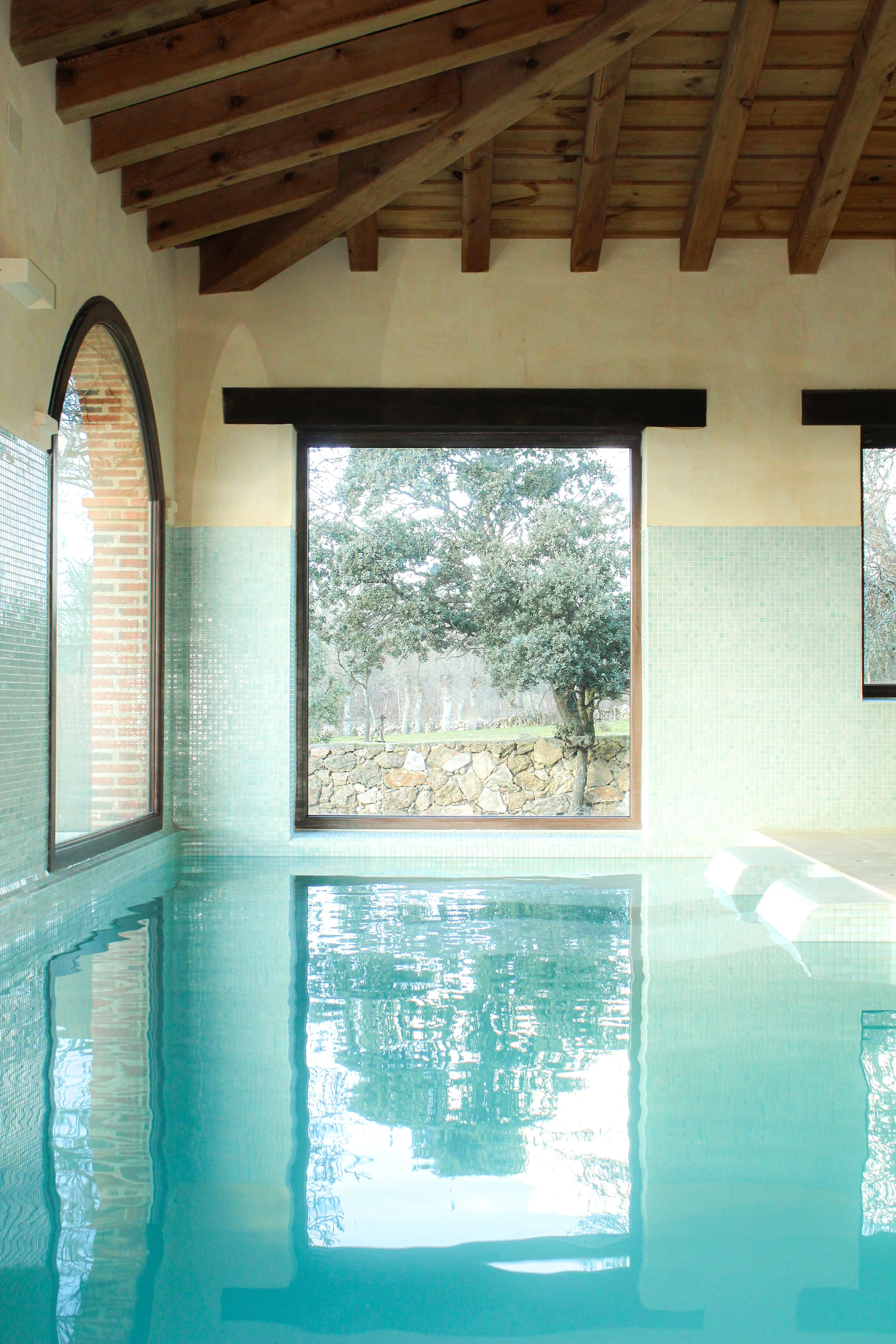 Piscina interior con ventanas grandes y vista a un paisaje con árboles y muro de piedra.
