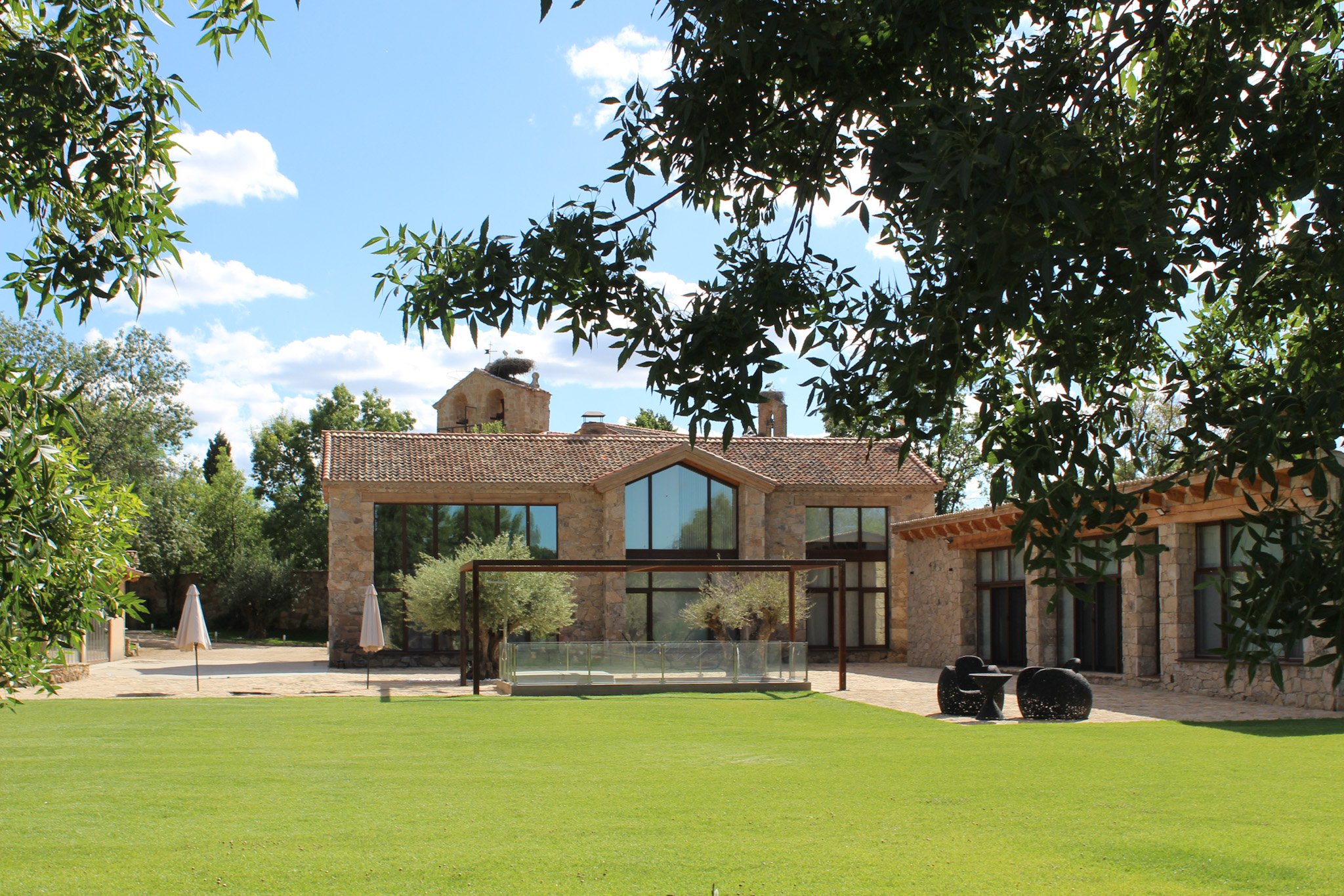 Casa de piedra con ventanas grandes, rodeada de un amplio césped verde y árboles, en un día soleado con algunas nubes en el cielo.