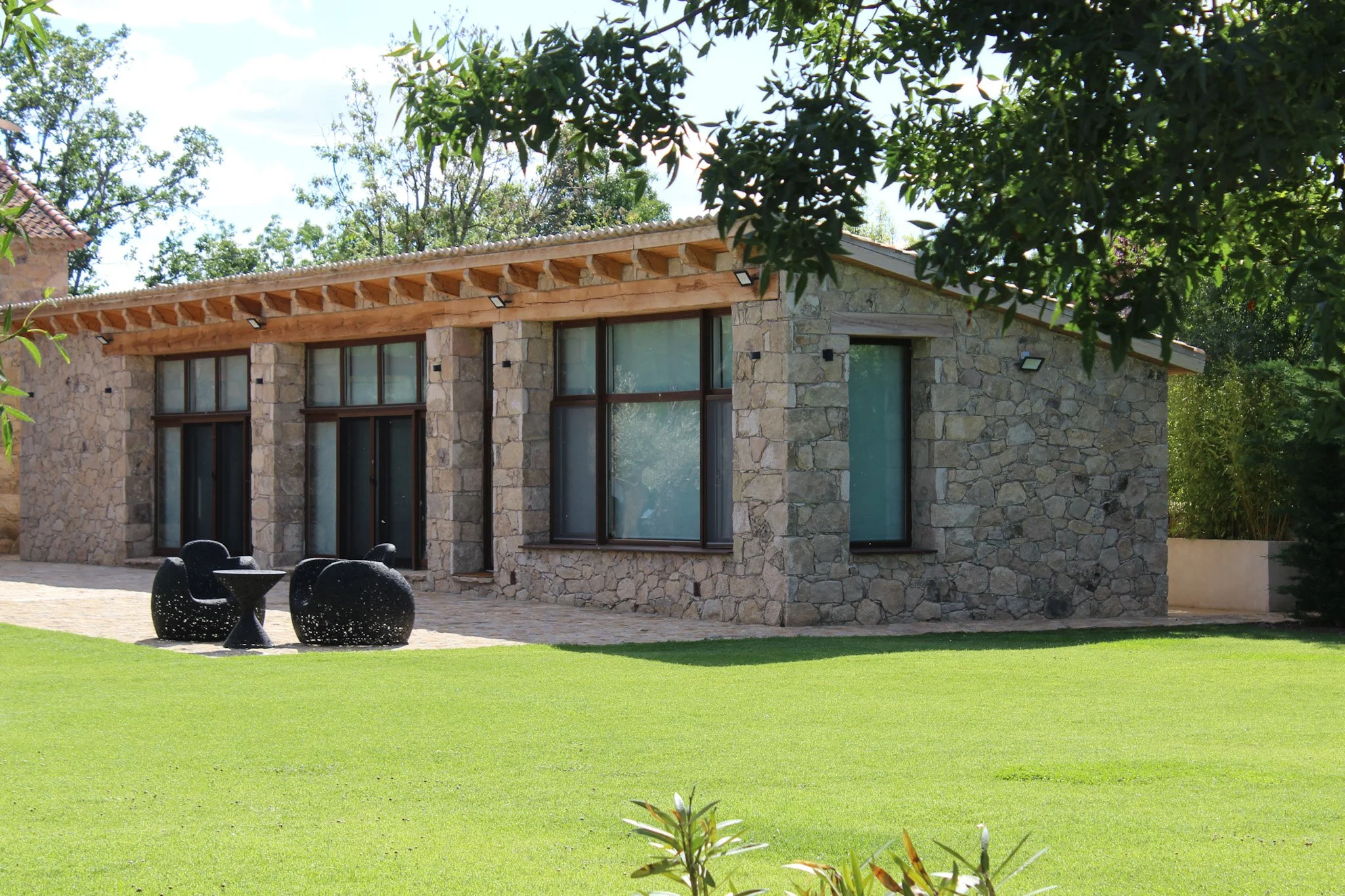 Casa moderna de piedra con grandes ventanas y un jardín de césped verde, con mobiliario de exterior negro.