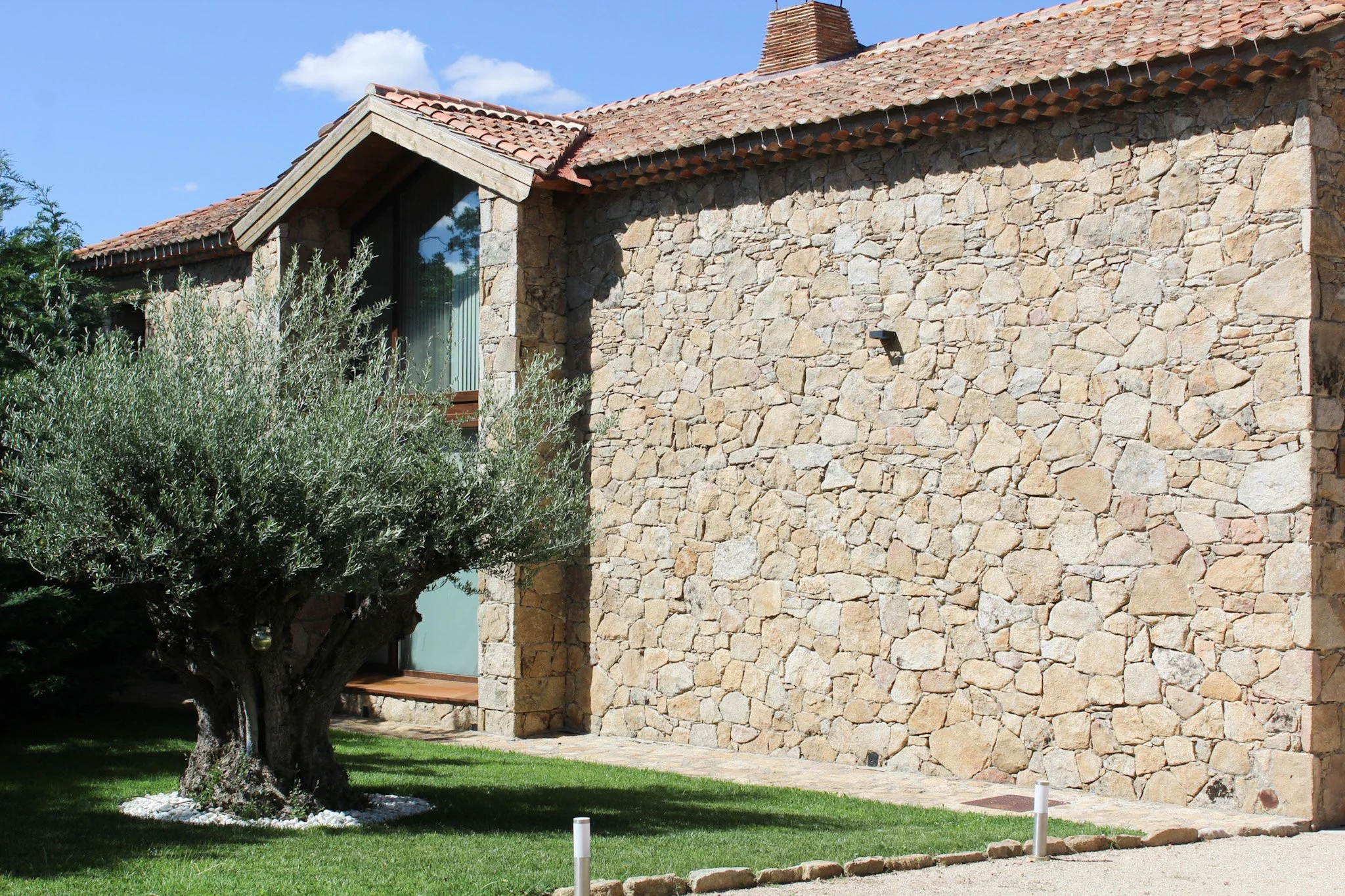 Casa de piedra con techo de tejas rojas, ventana grande y árbol en el jardín