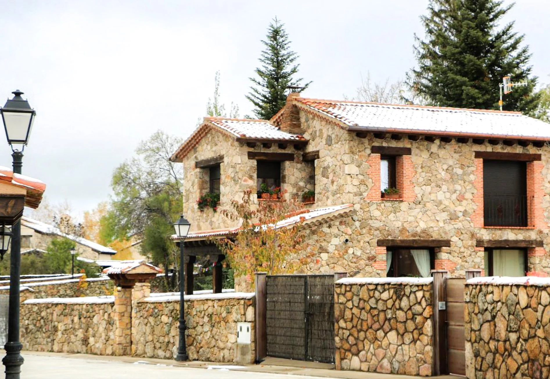 Casa de piedra con dos pisos, ventanas con macetas y cortinas, cerca de lámparas de calle, con un muro de piedra y un portón en la entrada, rodeada de árboles y nieve en el techo.