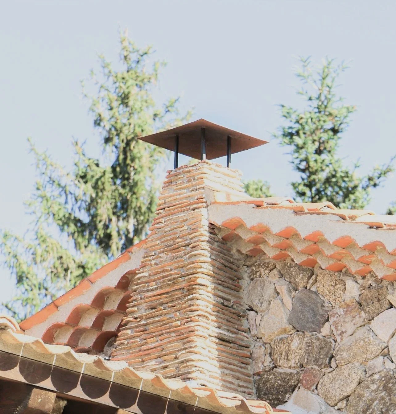 Chimenea de ladrillos en el techo de una casa rústica con tejas de barro y piedras, con árboles en el fondo.