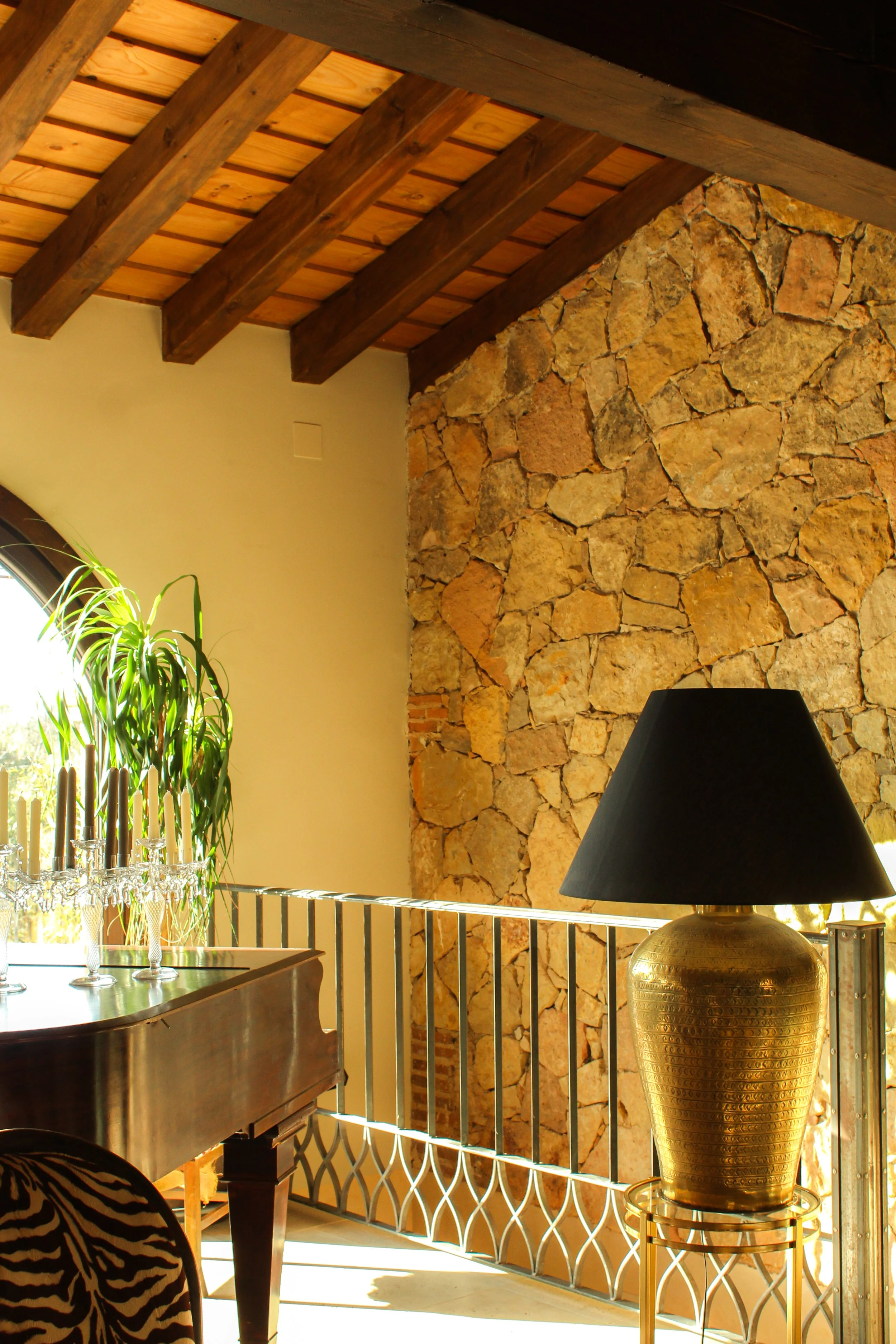 Interior de una casa con techo de vigas de madera, pared de piedra, una lámpara dorada y una planta junto a una ventana. Hay también un mueble de piano y varias copas de cristal.
