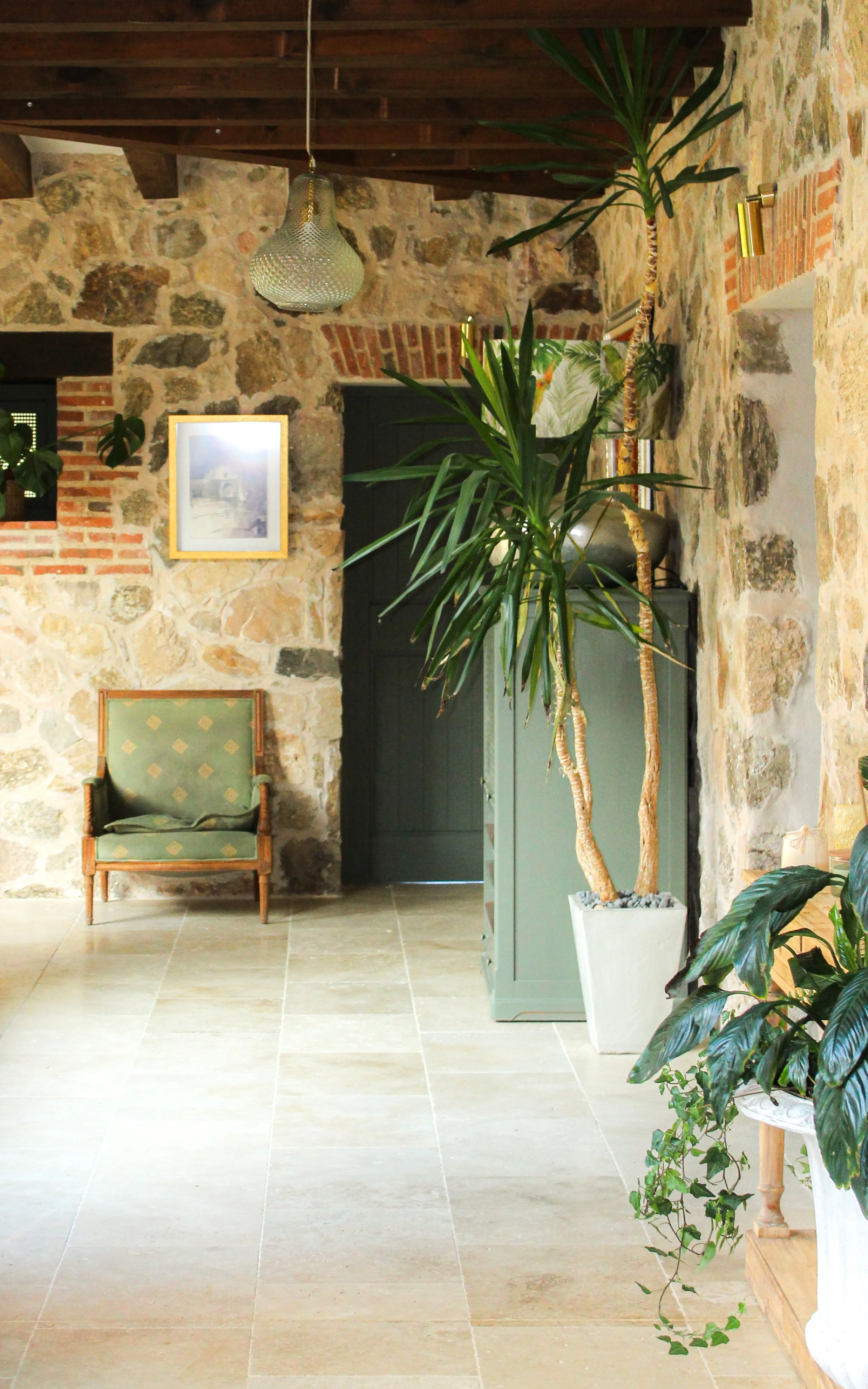 Interior de una habitación rústica con paredes de piedra, un armario gris, un sillón verde y plantas grandes decorativas.