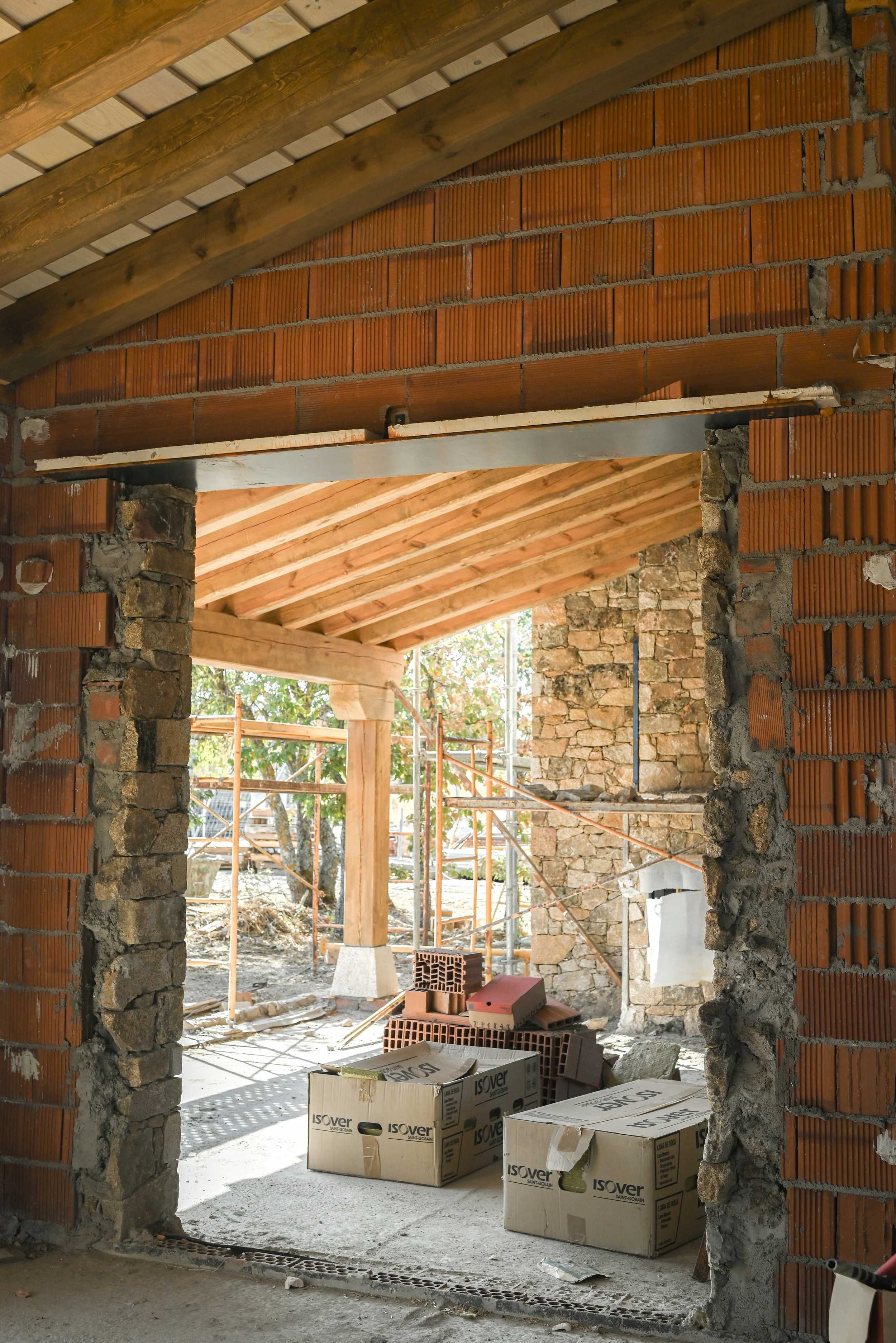 Construcción de una casa con paredes de ladrillo, techo de madera y ventanas abiertas, con materiales de construcción y herramientas en el interior.