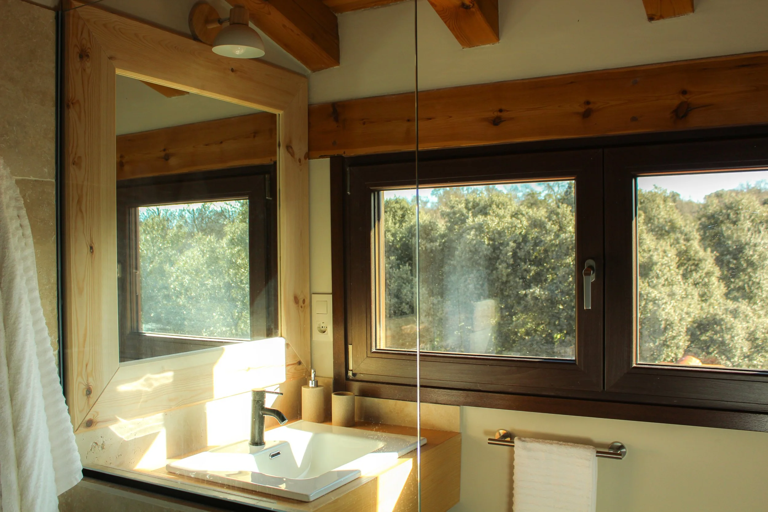 Cuarto de baño con ventanas grandes de madera, lavabo con grifo negro, espejo y toalla en soporte.