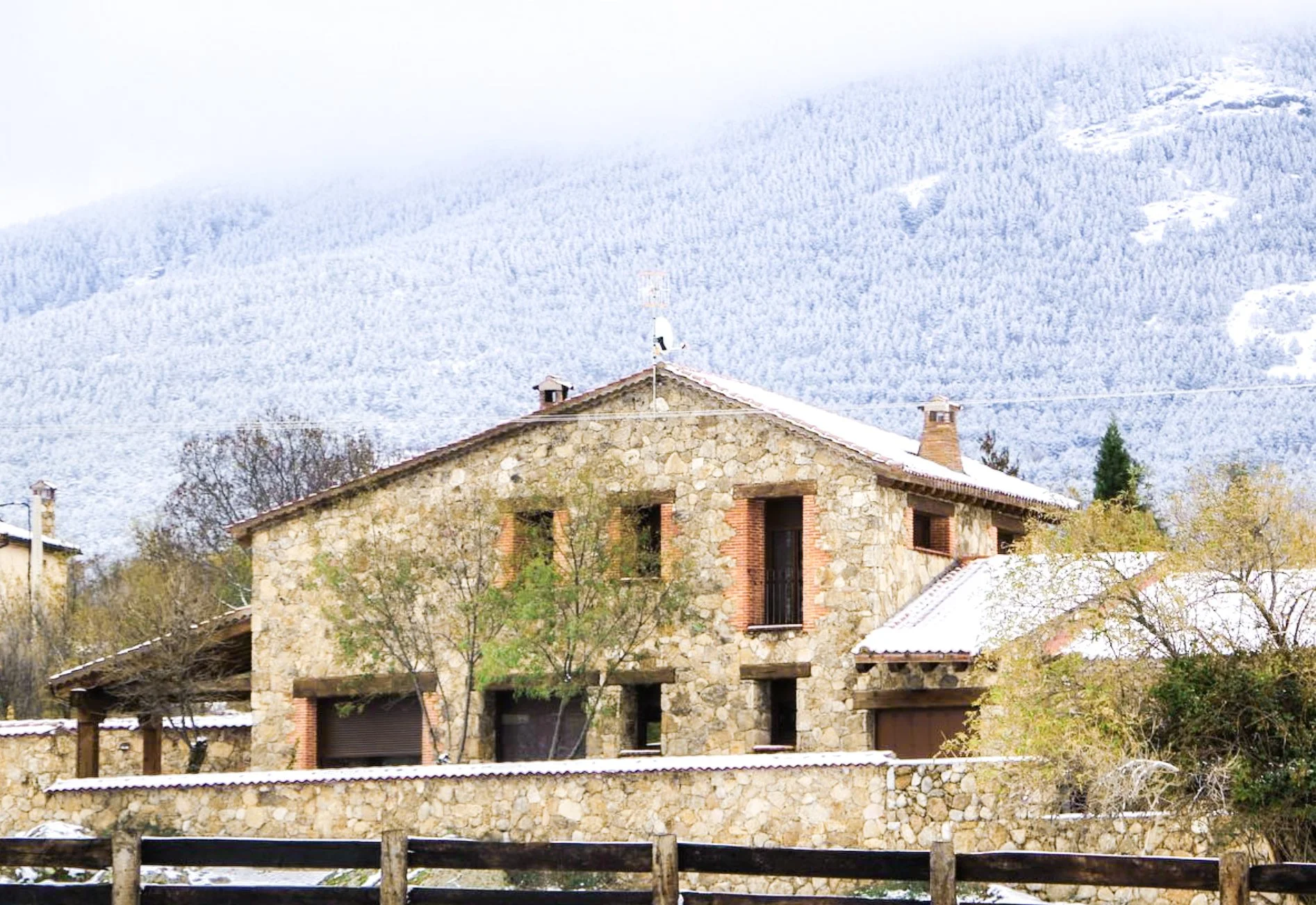 Casa de piedra con tejado cubierto de nieve, árboles y montañas nevadas en el fondo.