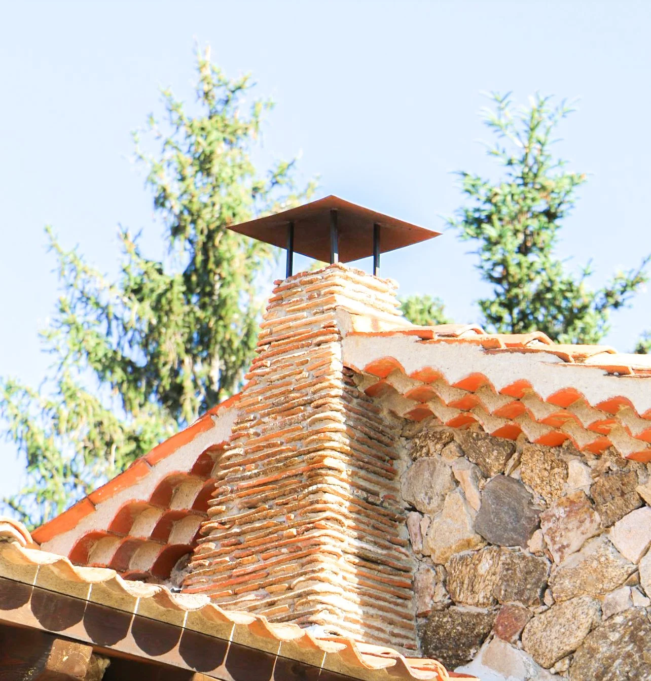 Chimenea de ladrillos en la azotea de una casa de piedra con tejas de cerámica, con árboles verdes en fondo y cielo azul.
