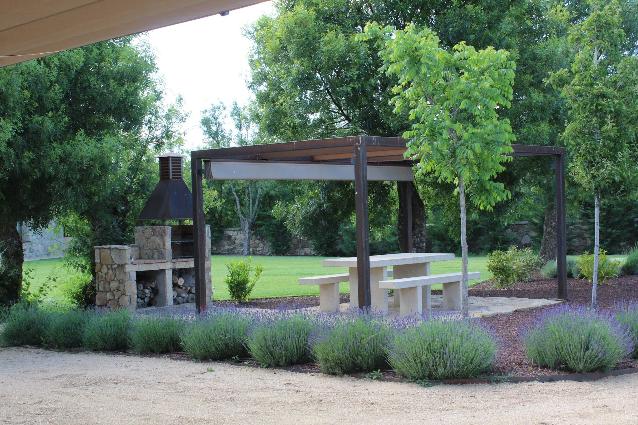 Área de descanso con mesa de concreto, bancos, estructura de sombra, una parrilla de piedra y un jardín de lavanda en primer plano, rodeado de árboles y césped