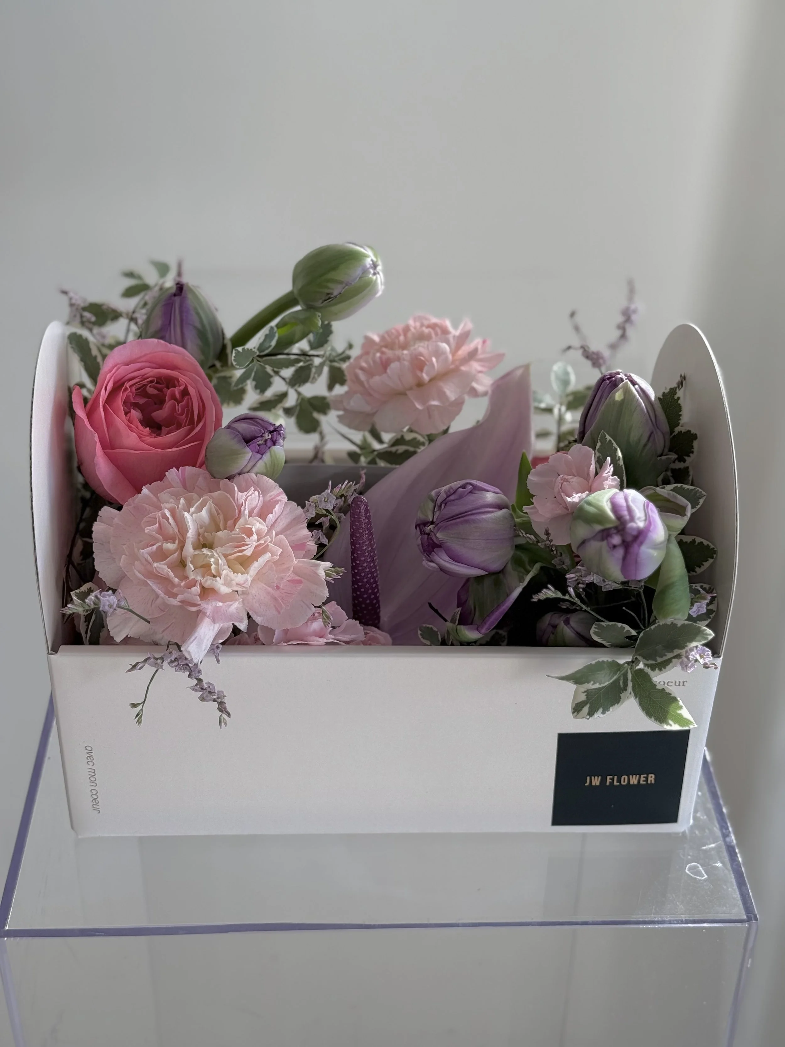 A white box filled with pink and purple flowers, including roses and carnations, arranged with greenery.