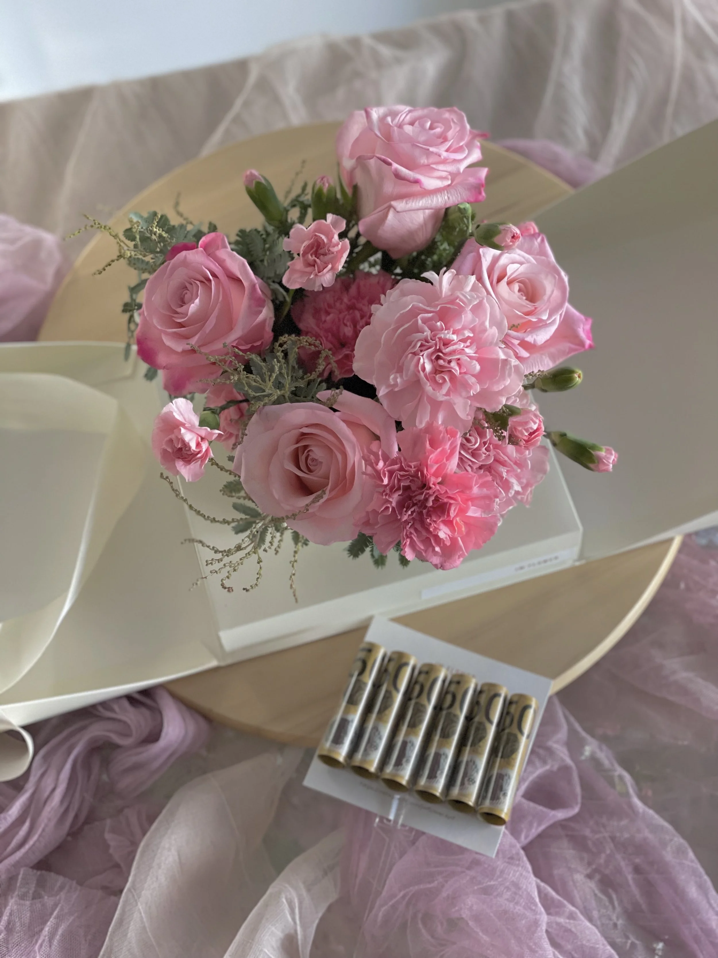 A bouquet of pink roses, carnations, and greenery in a white box, placed on a wooden table surrounded by pink and beige fabric.