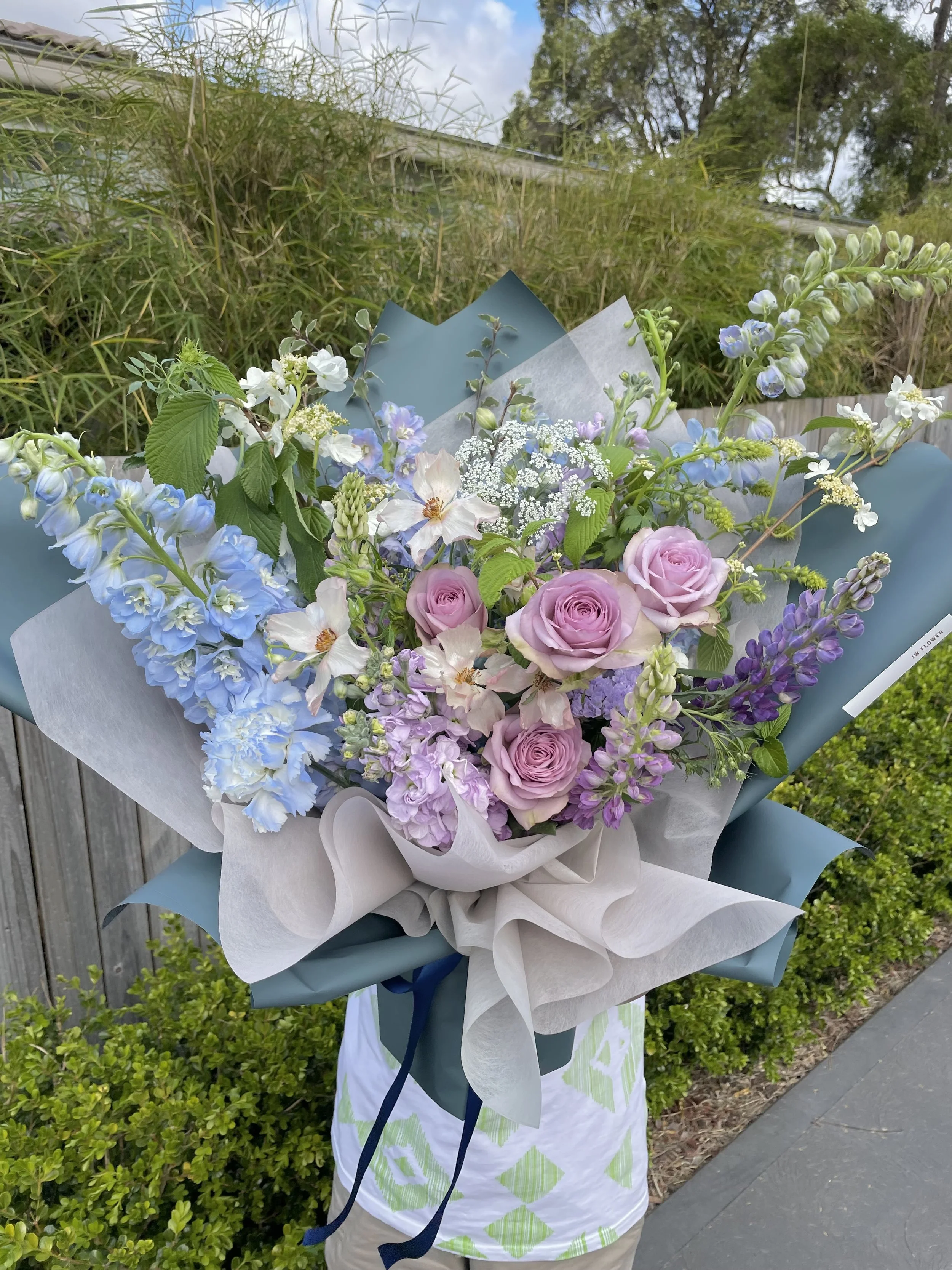 A floral bouquet featuring lavender roses, delphiniums, and other small pastel-colored flowers, wrapped in decorative paper and held outdoors near a fence and greenery.