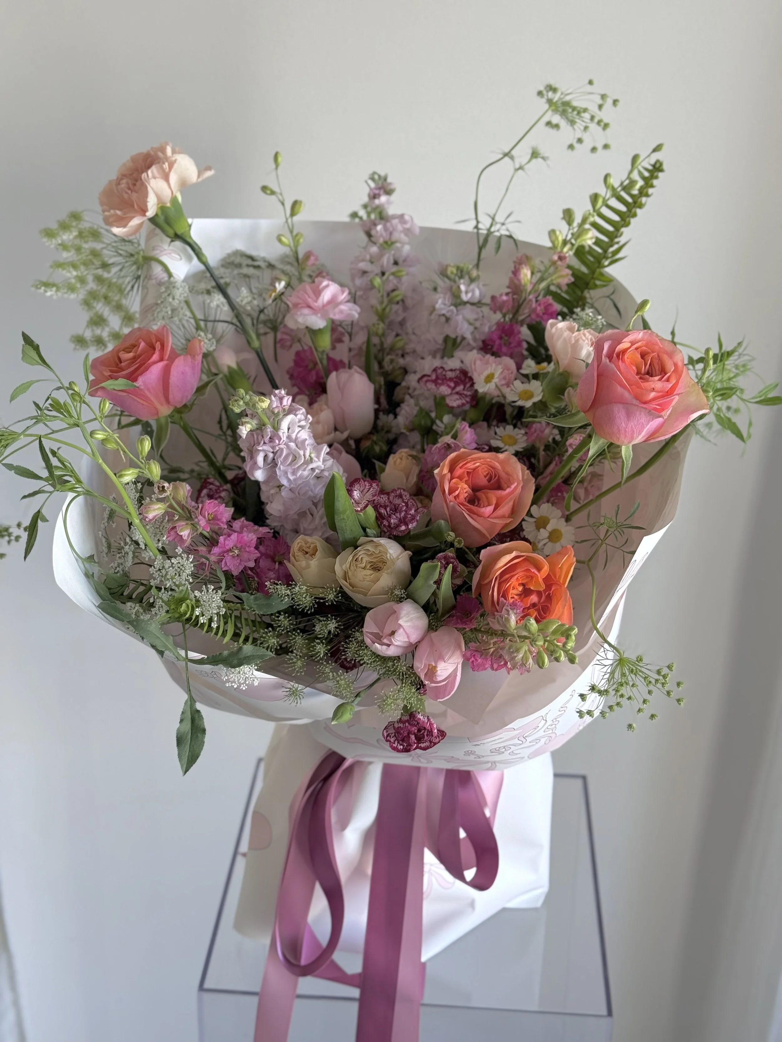 A colorful bouquet of mixed pink, peach, and cream flowers with green foliage wrapped in white and pink paper, tied with a pink ribbon.