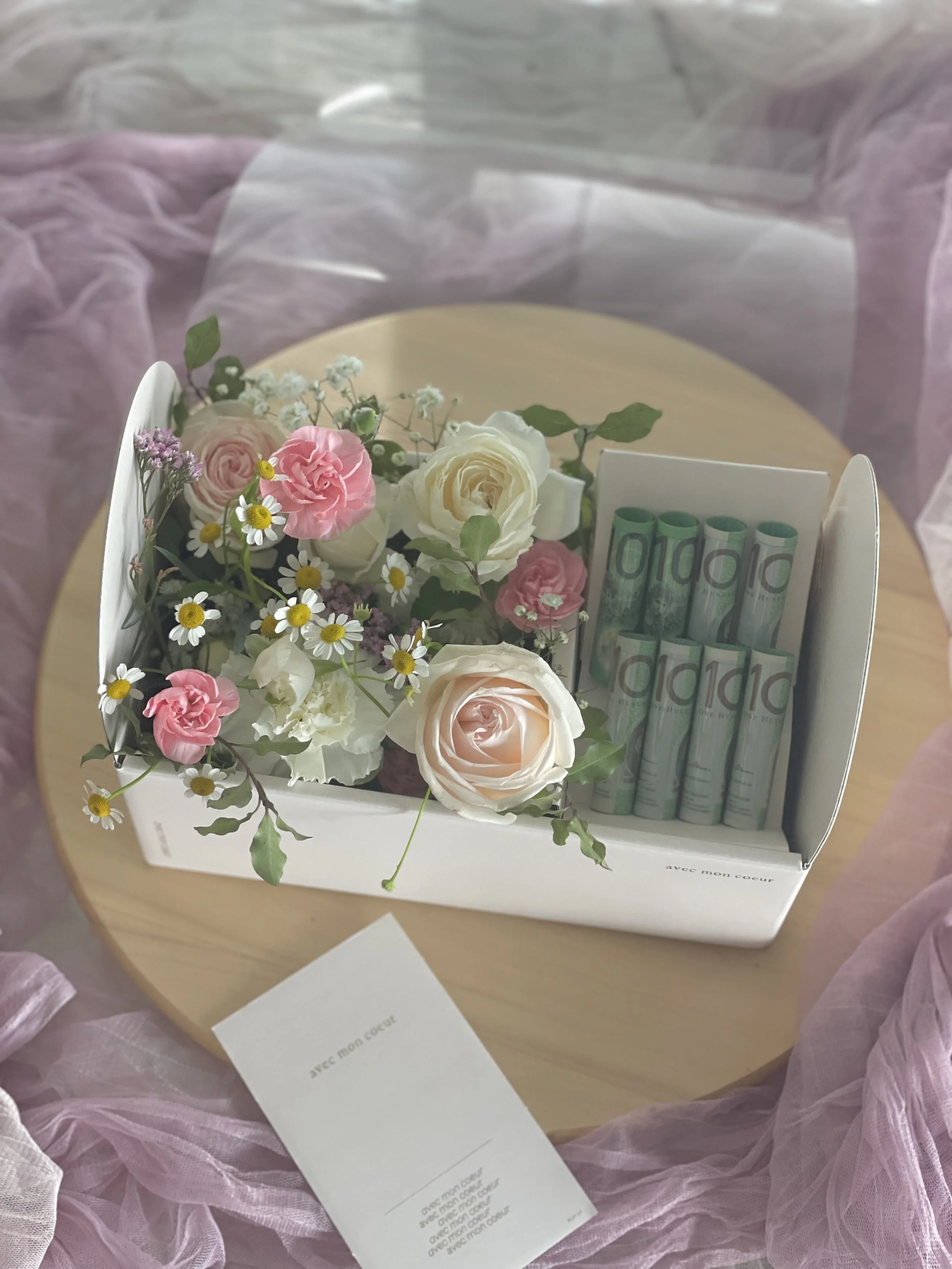 A gift box with a floral arrangement of pink and white roses, daisies, and other flowers, alongside a box of rolled-up greeting cards or scrolls, placed on a wooden surface with pink tulle fabric surrounding it.