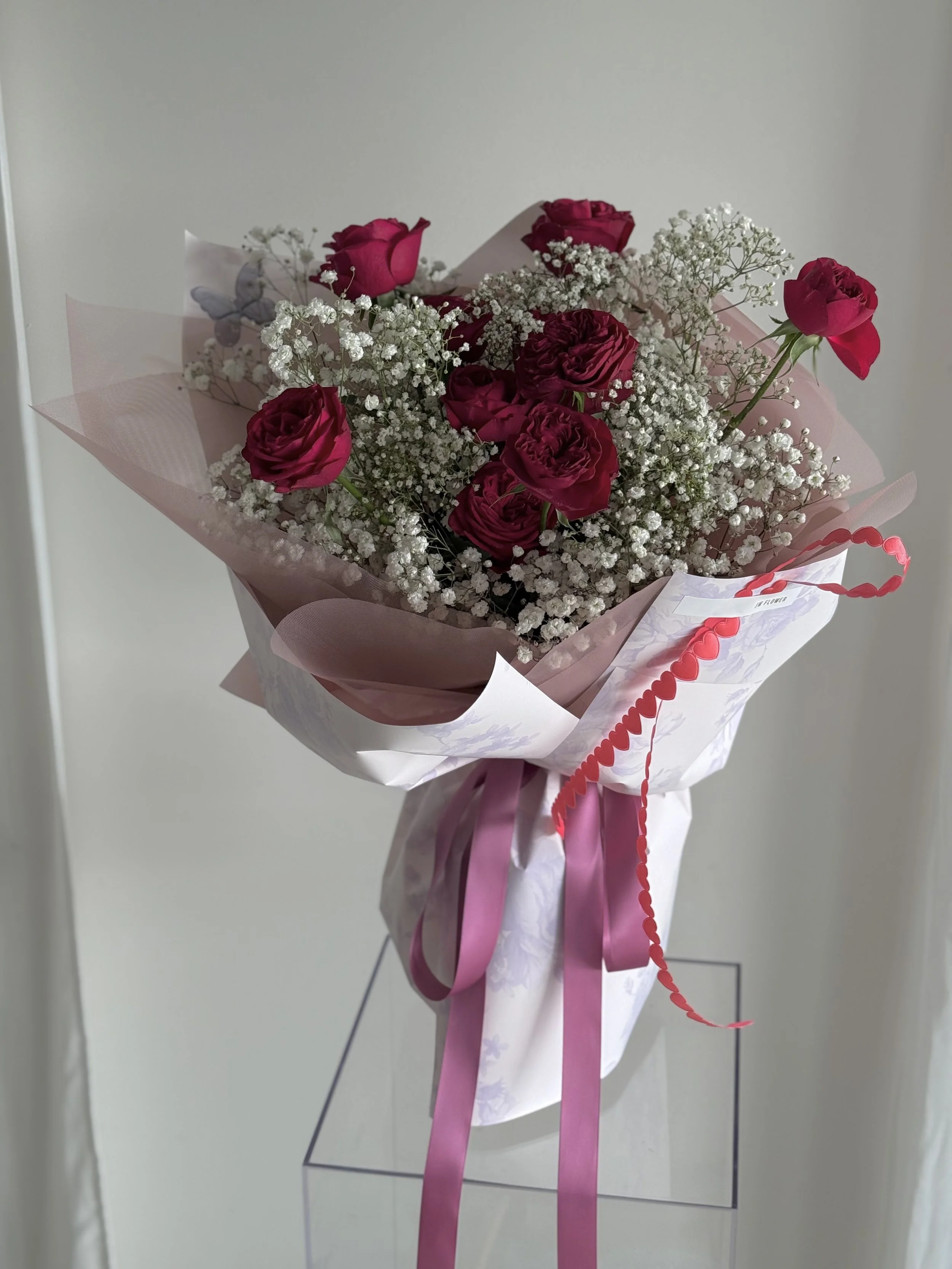 A bouquet of pink roses and white baby's breath flowers wrapped in pink and white paper, decorated with pink ribbons and a red heart-shaped string.