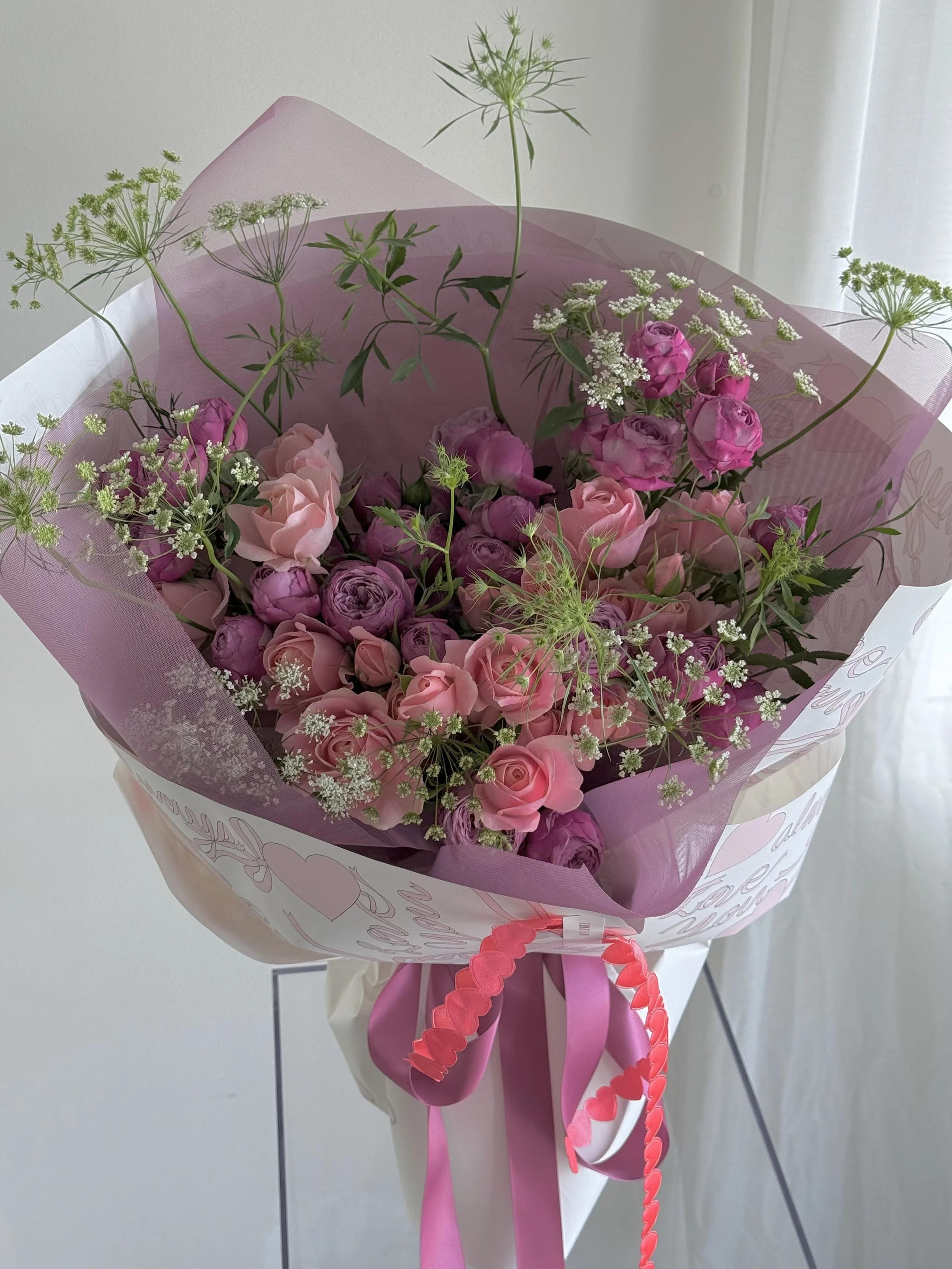 A bouquet of pink and purple roses wrapped in pink and purple tissue paper with ribbon, decorated with white baby's breath and delicate greenery.