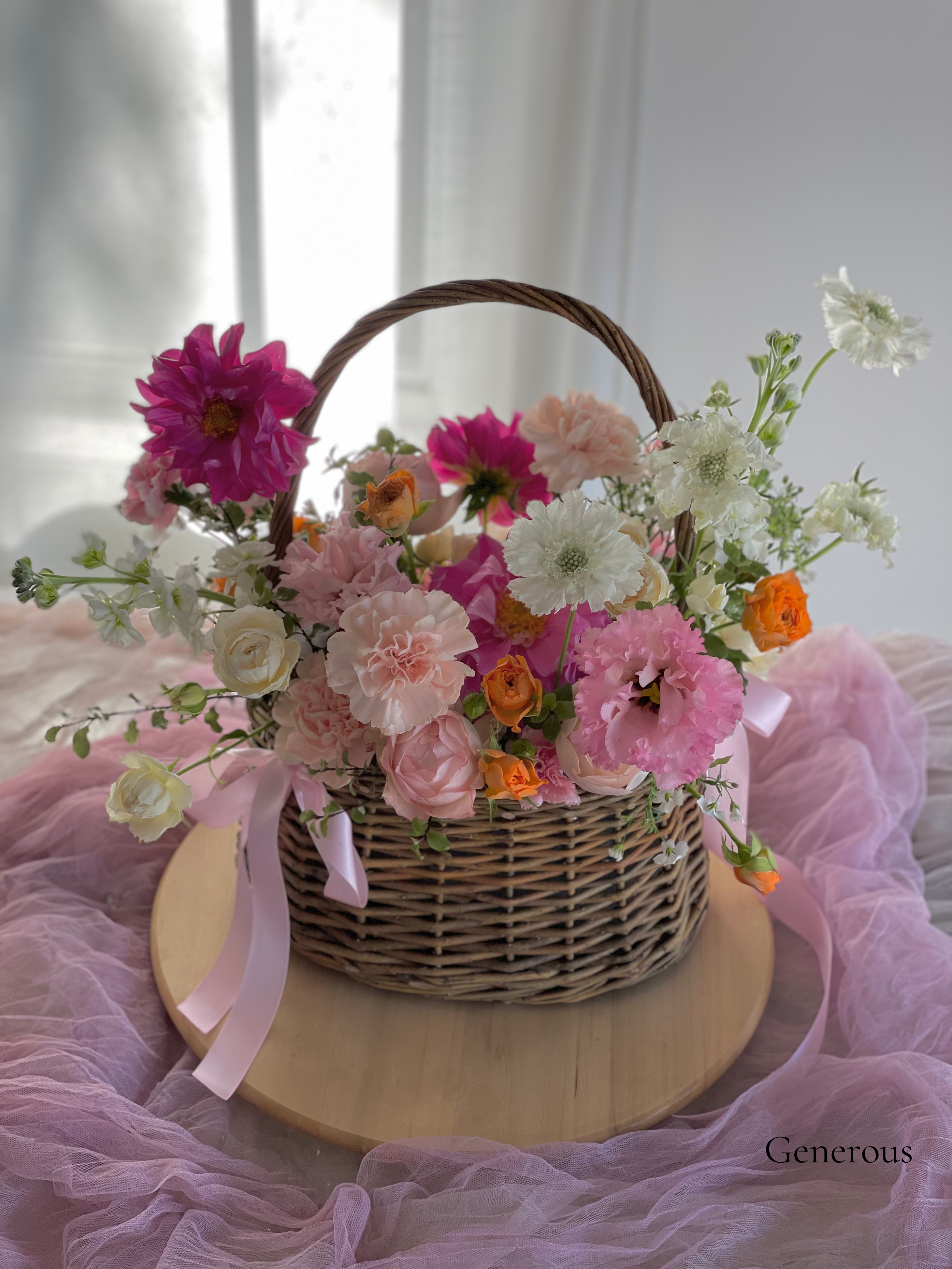 A wicker basket filled with pink, white, orange, and purple flowers resting on a round wooden platform, with pink tulle fabric beneath, near a window.