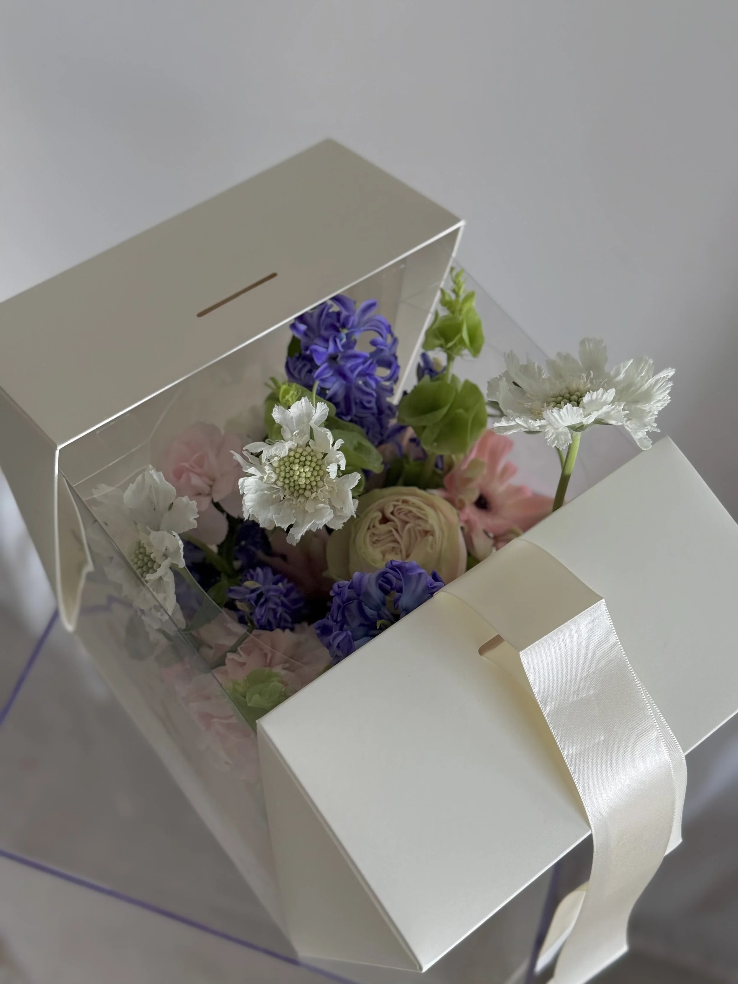 A white gift box with a satin ribbon containing a colorful bouquet of flowers, visible through a see-through window.
