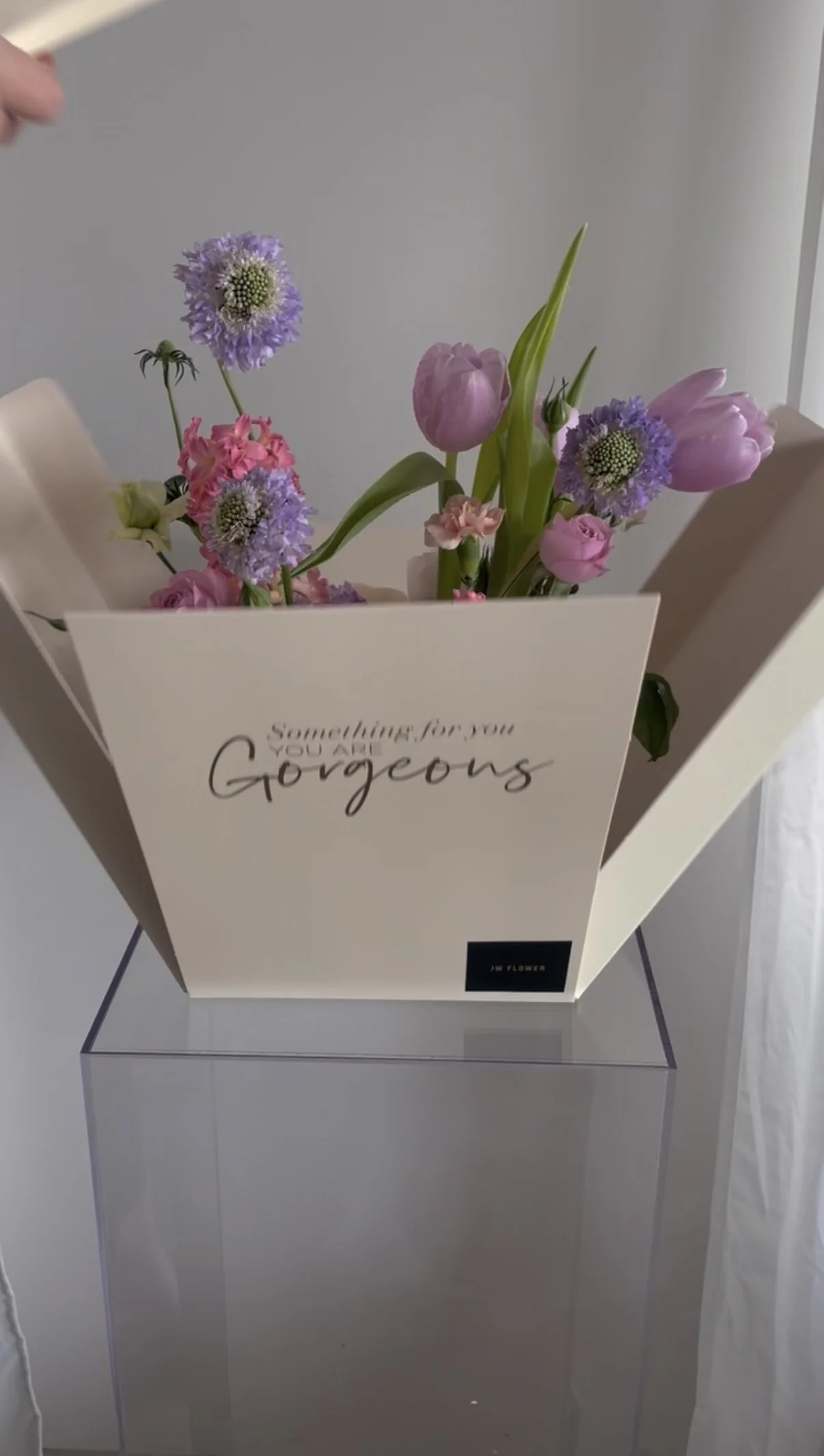 A bouquet of pink and purple flowers in a white box with the words "Something for you, you are gorgeous" written on it, placed on a clear display stand.