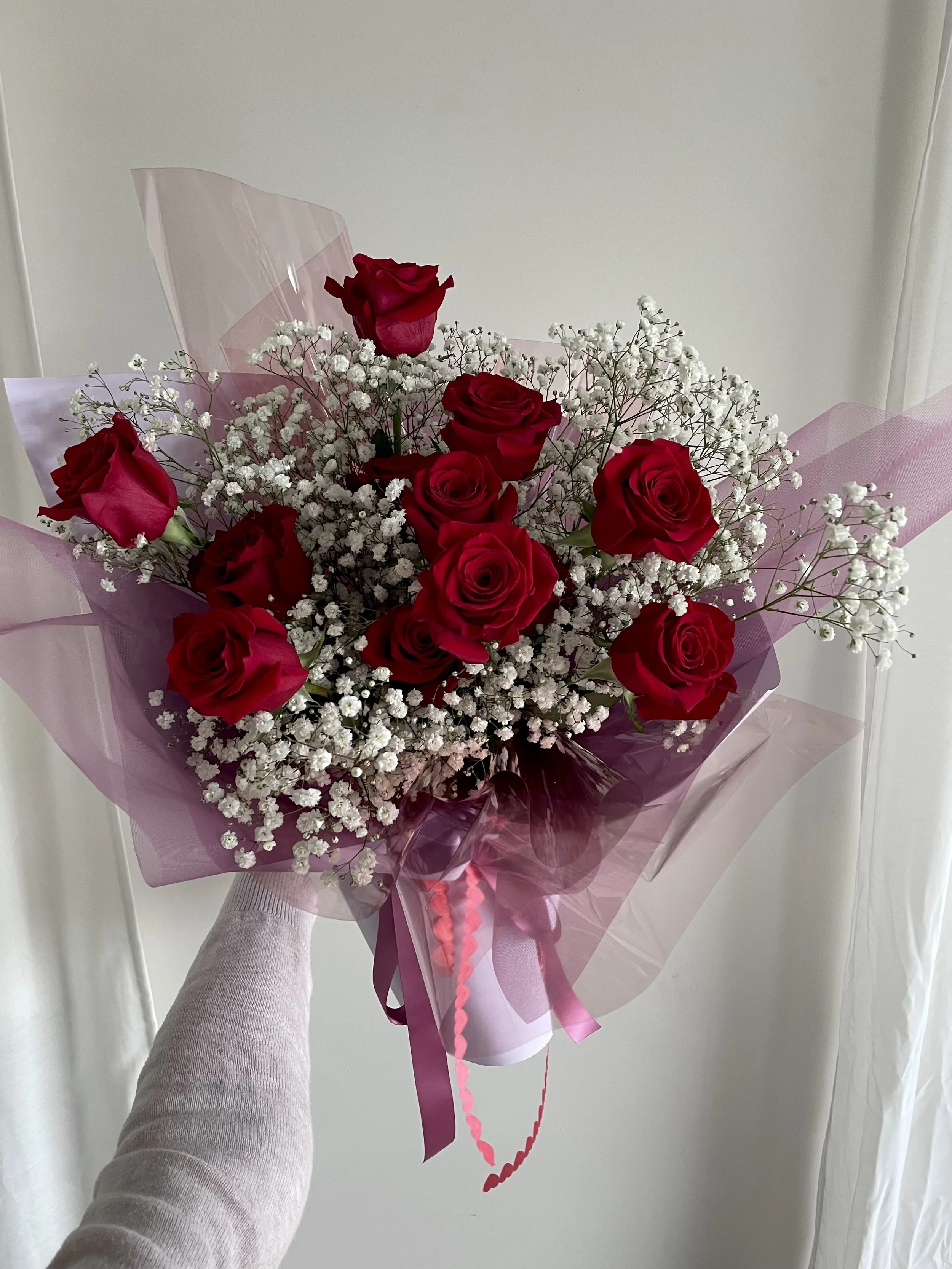 A bouquet of red roses and baby's breath flowers wrapped in pink and clear cellophane with pink and red ribbons being held by a person wearing a light-colored long sleeve shirt, against a plain light-colored background.